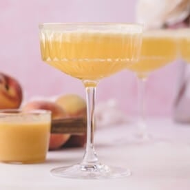 A coupe glass filled with a light orange cocktail, with peaches and another glass in the background on a light pink surface.