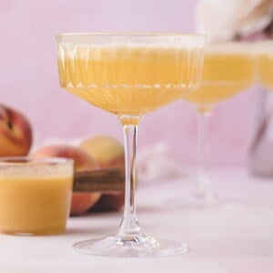 A coupe glass filled with a light orange cocktail, with peaches and another glass in the background on a light pink surface.