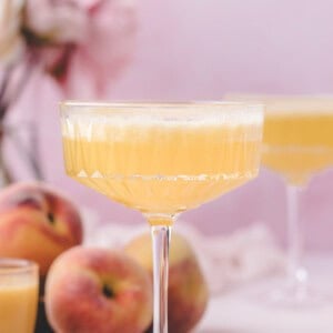 A coupe glass filled with a light orange peach bellini is in focus, with fresh peaches and a blurred pink floral arrangement in the background.