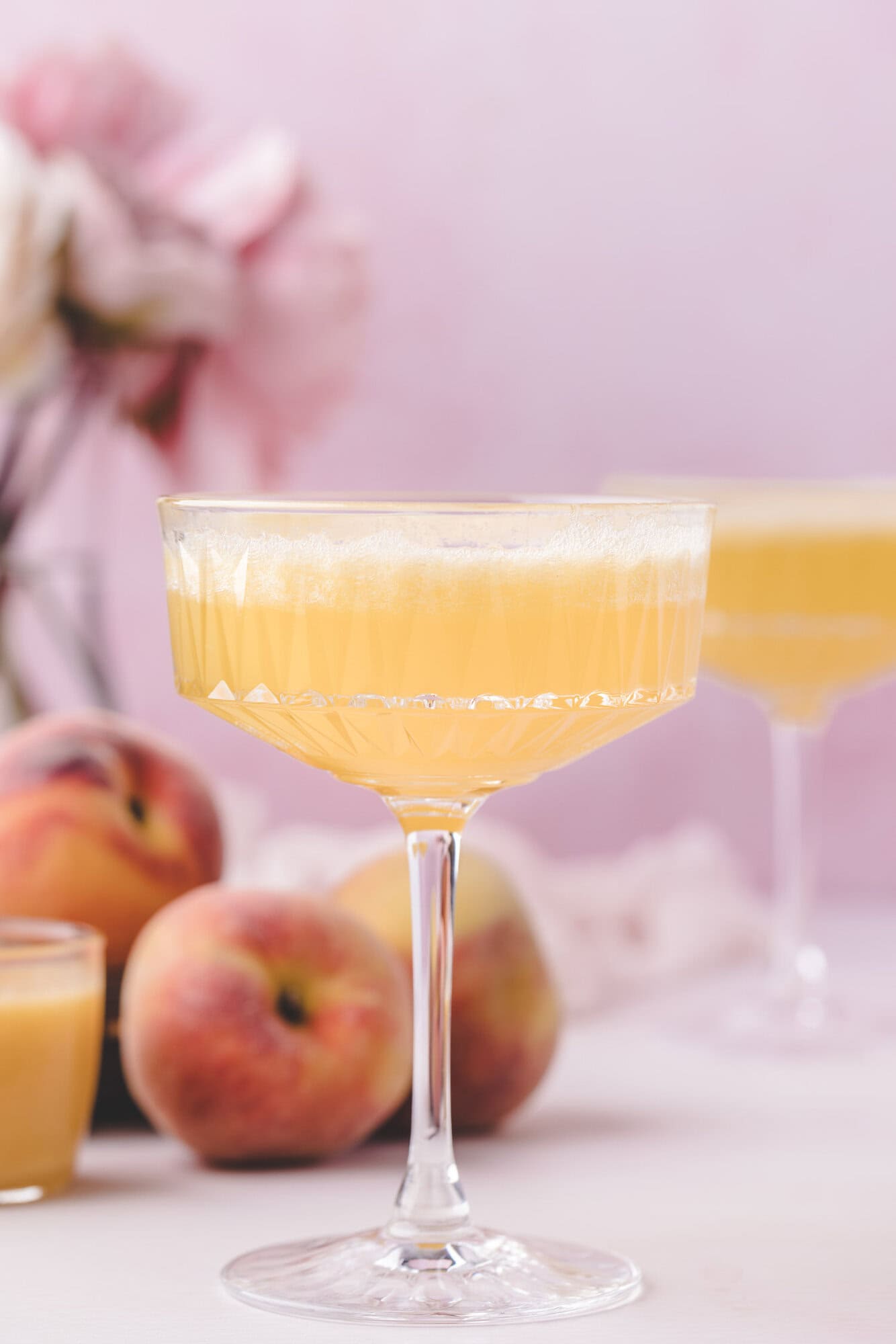 A coupe glass filled with a light orange peach bellini is in focus, with fresh peaches and a blurred pink floral arrangement in the background.