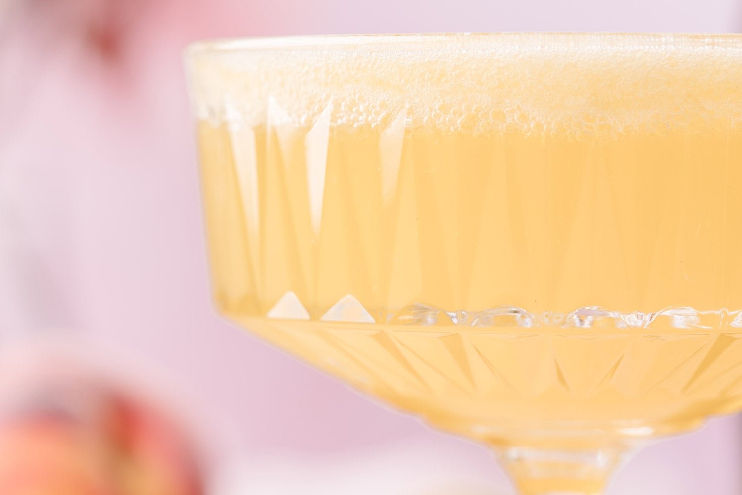 Close-up of a coupe glass filled with a light yellow, frothy beverage against a soft, blurred background.