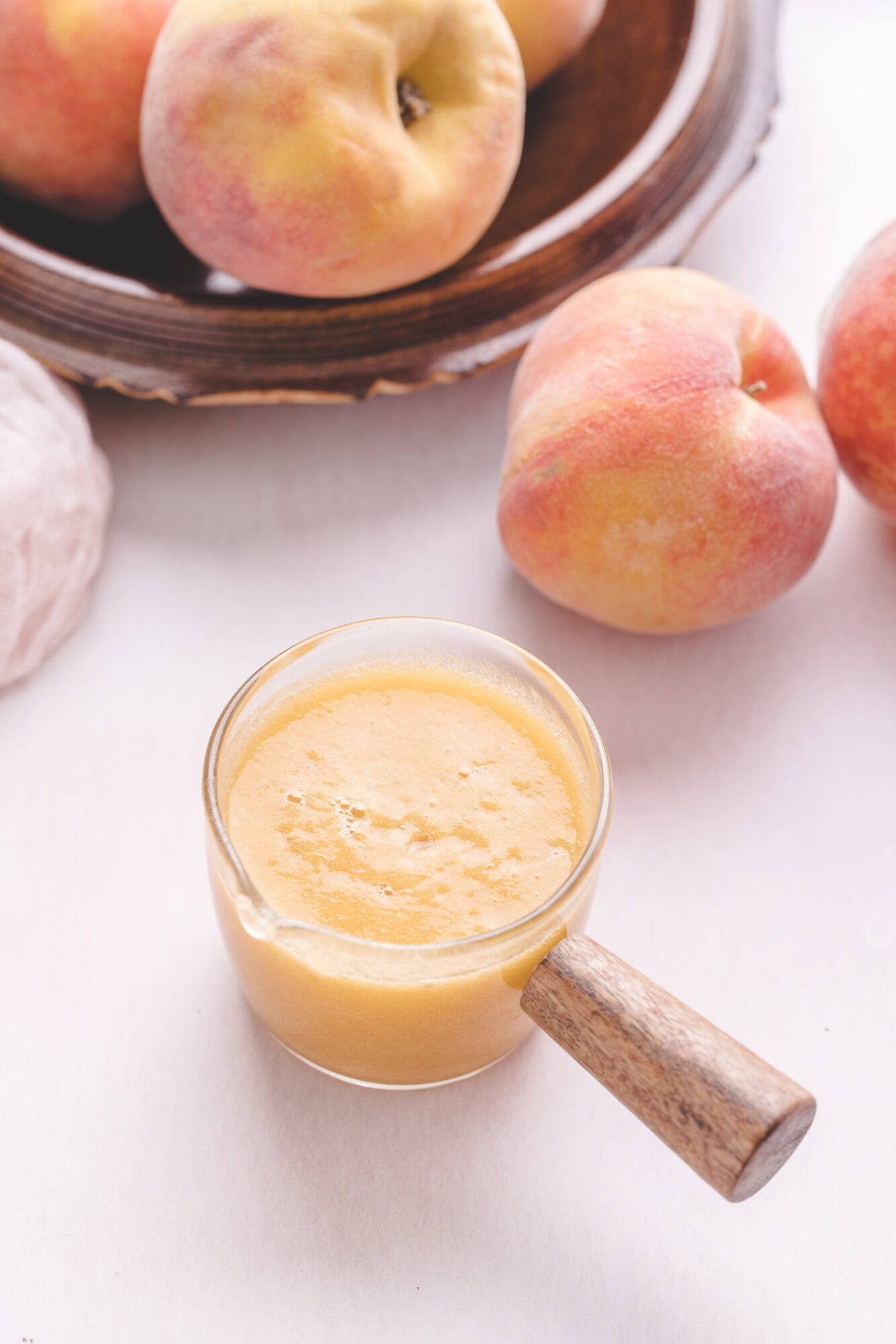 A glass container filled with peach smoothie sits on a white surface, with whole peaches and a wooden plate in the background.
