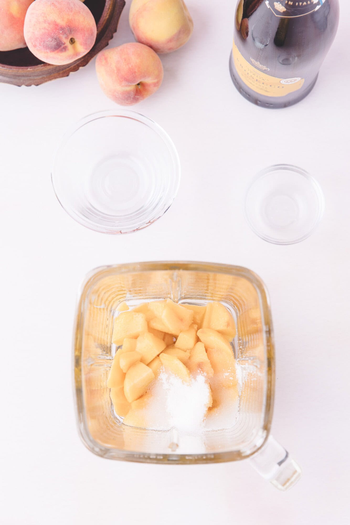 A blender contains chopped peaches and sugar. Nearby are empty glass bowls, a bottle of sparkling wine, and a bowl of whole peaches on a white surface.