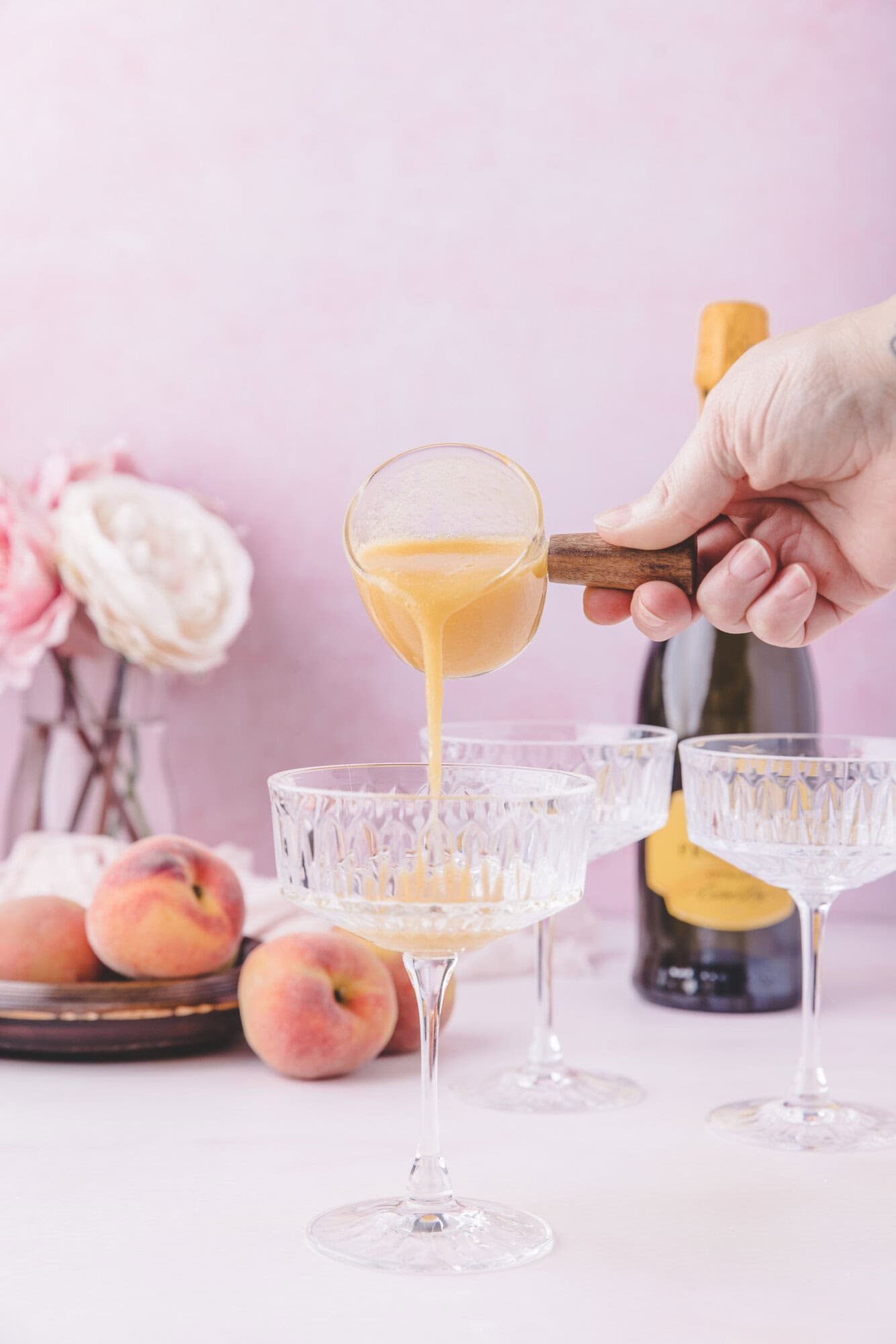 A hand pours peach puree into a coupe glass, surrounded by fresh peaches, a bottle of sparkling wine, and additional empty glasses on a pink background.