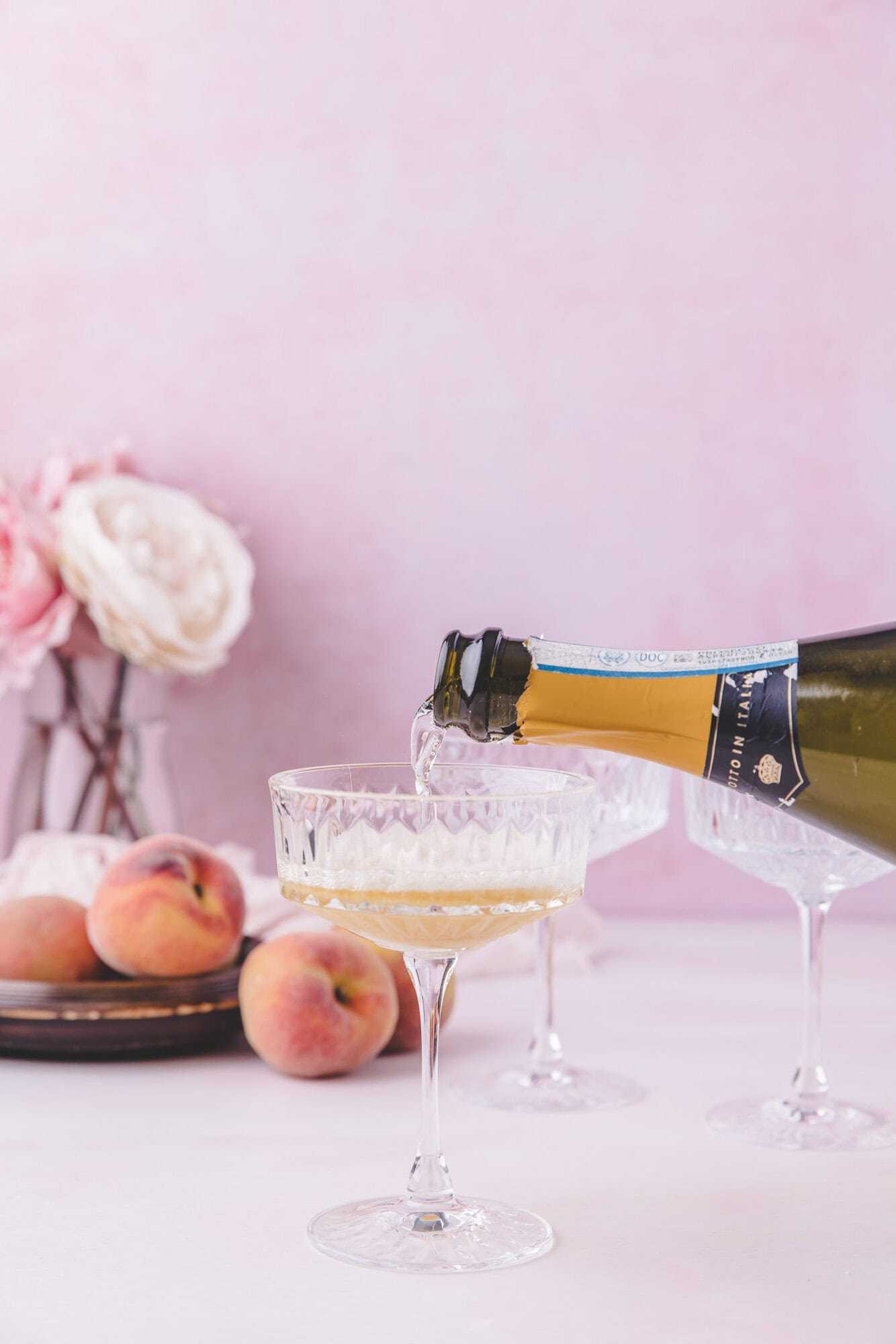 A bottle of sparkling wine is being poured into a coupe glass, with peaches and flowers arranged on a table in the background.