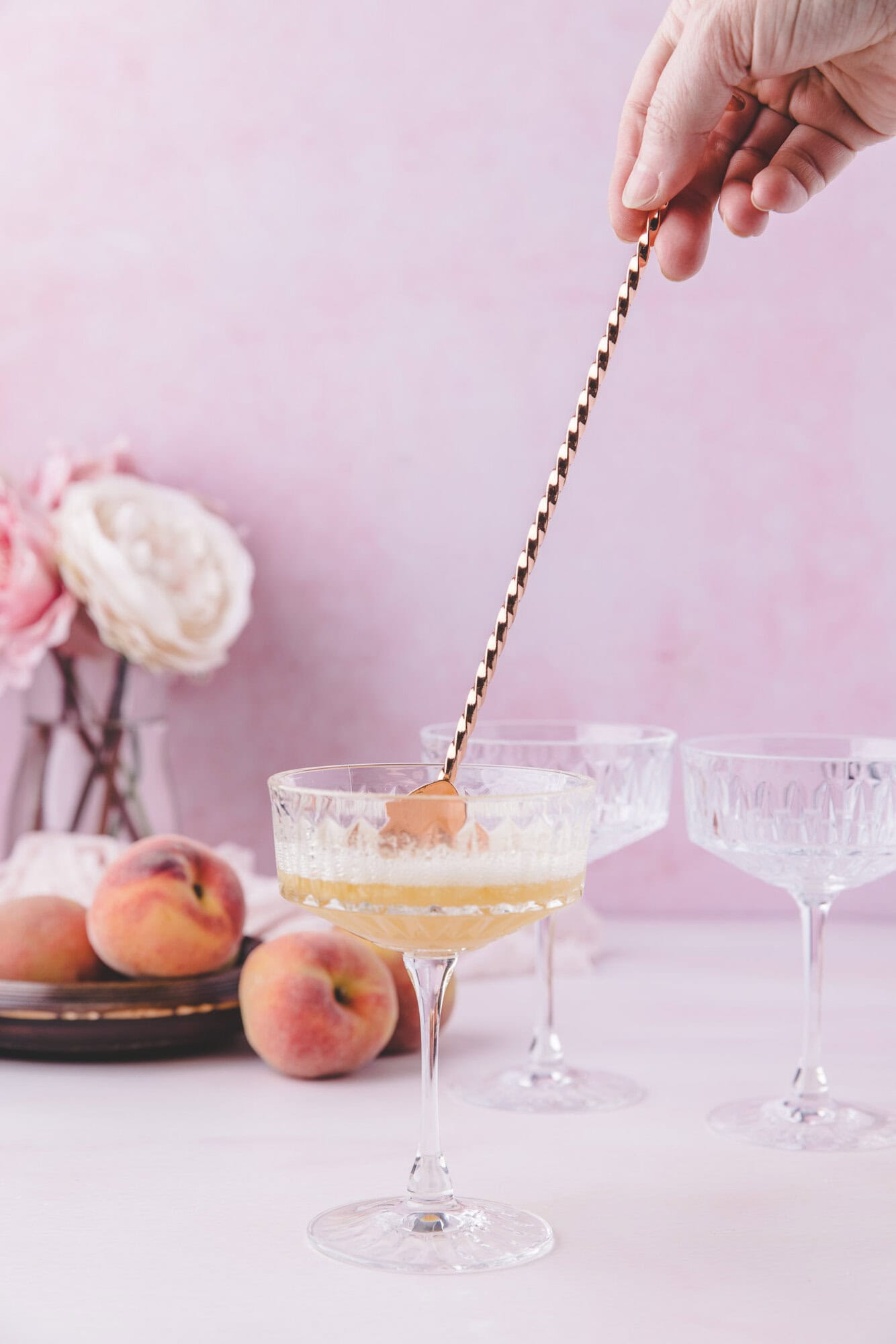 A hand stirs a drink in a coupe glass with a long spoon; peaches and flowers are arranged on a plate in the background.