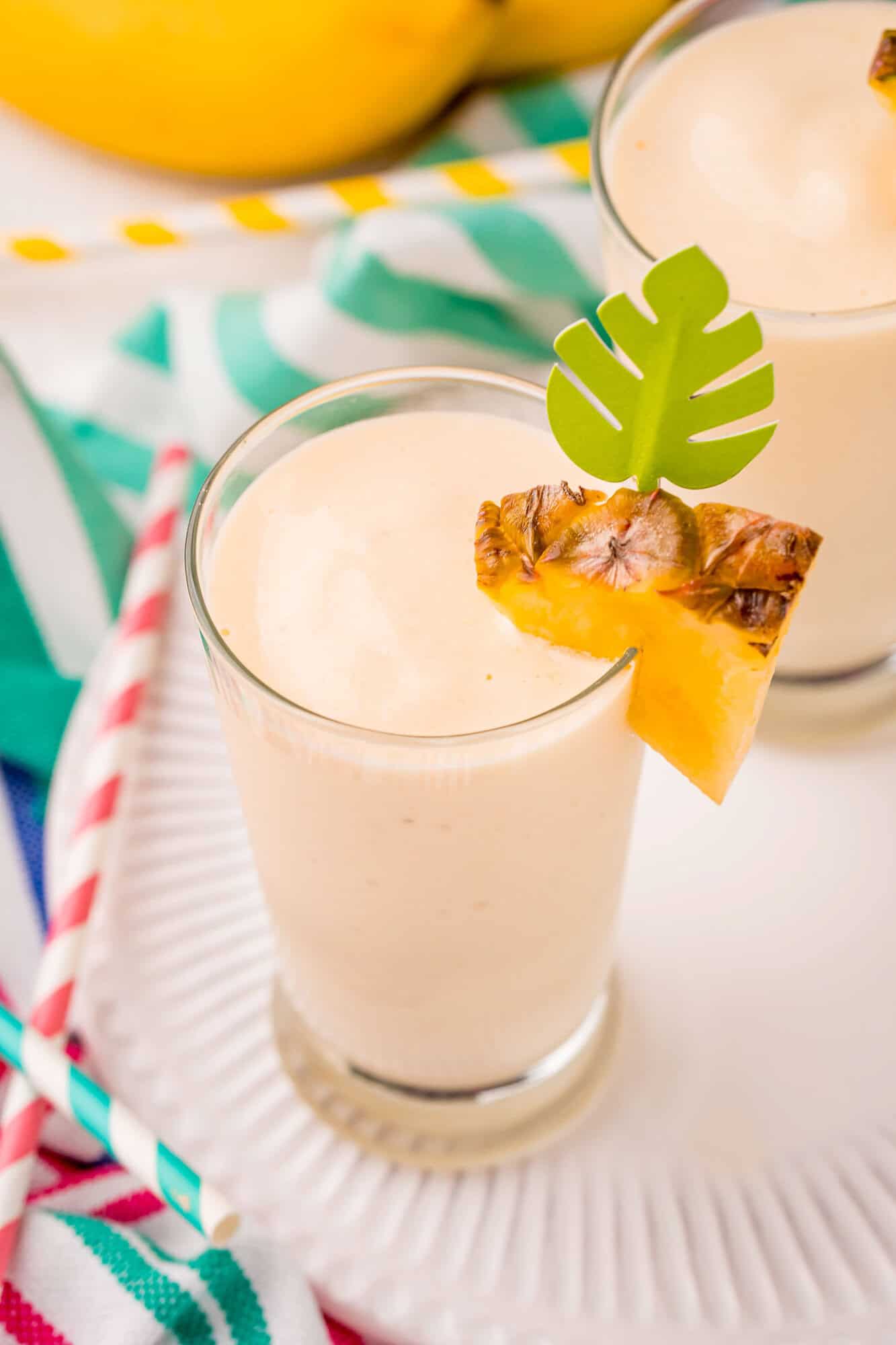 A glass of creamy smoothie garnished with a pineapple slice and a green paper leaf, placed on a white plate with colorful straws nearby.