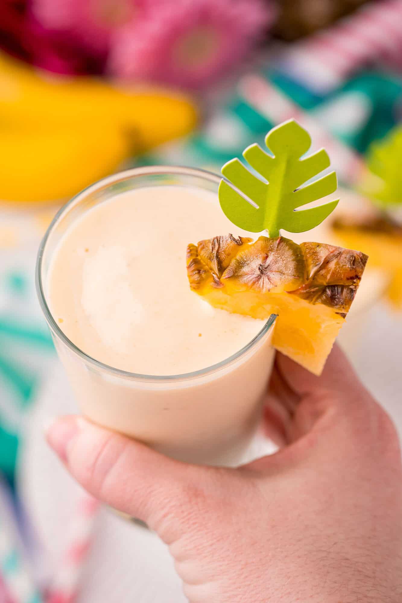 A hand holds a glass of creamy smoothie garnished with a pineapple slice and a decorative green paper leaf.