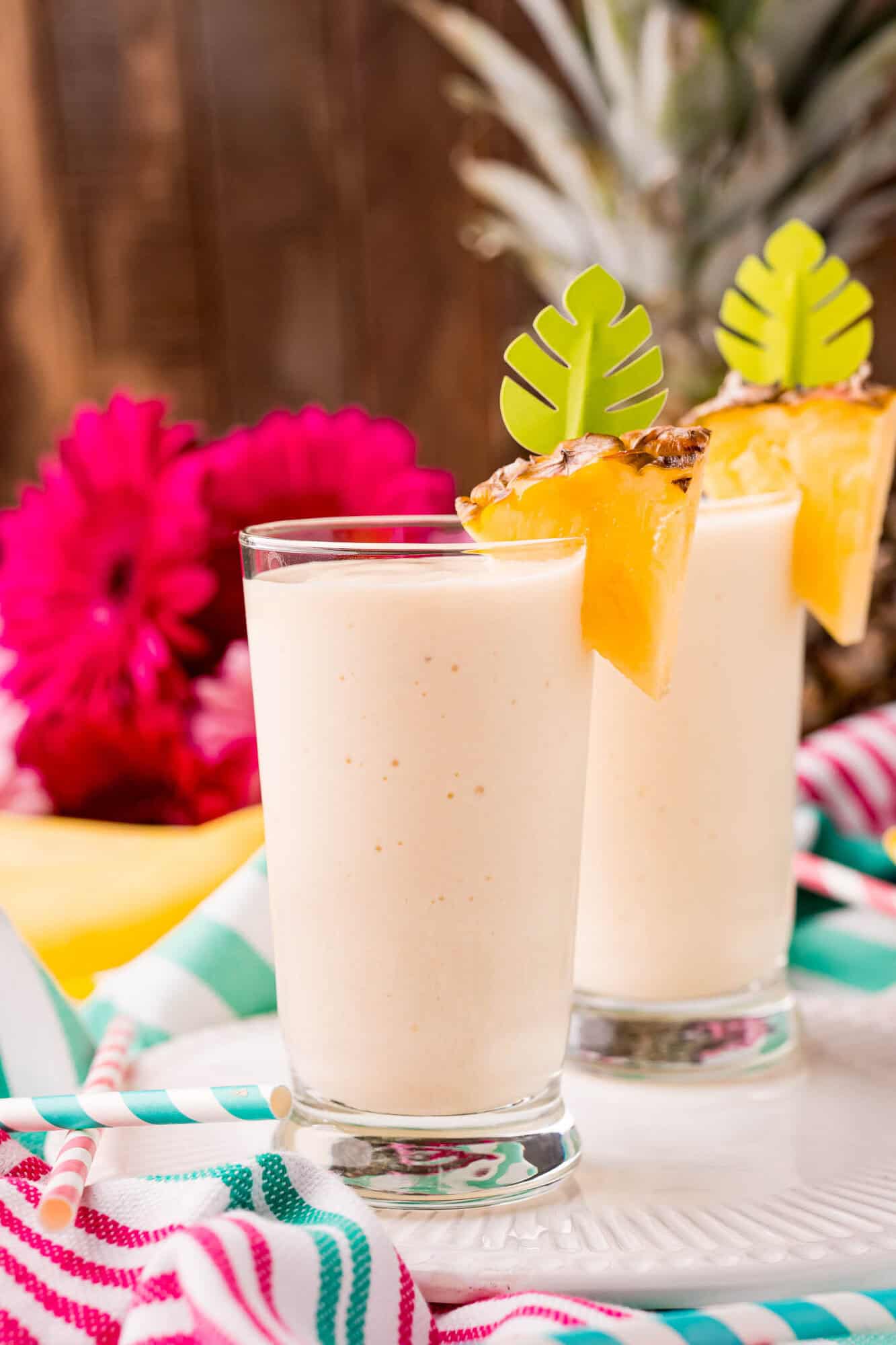 Two glasses of creamy pineapple smoothie garnished with pineapple wedges and green leaf stirrers, with a colorful striped cloth and pink flowers in the background.