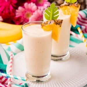 Two glasses of creamy smoothie garnished with pineapple slices and decorative leaf picks, placed on a white plate with striped straws nearby; flowers and a banana in the background.