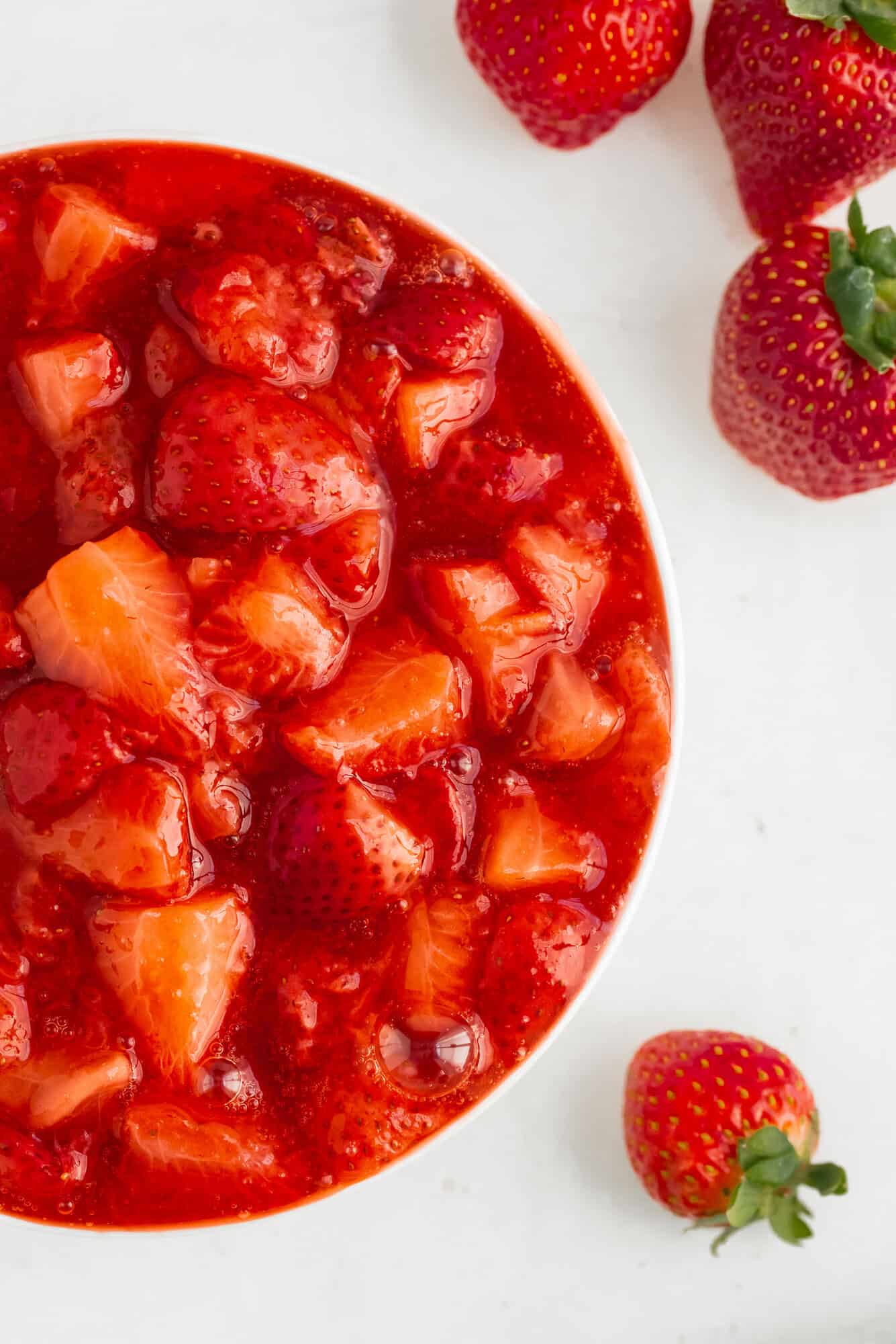 A bowl of strawberry compote with visible fruit chunks, placed on a white surface, with whole strawberries beside it.