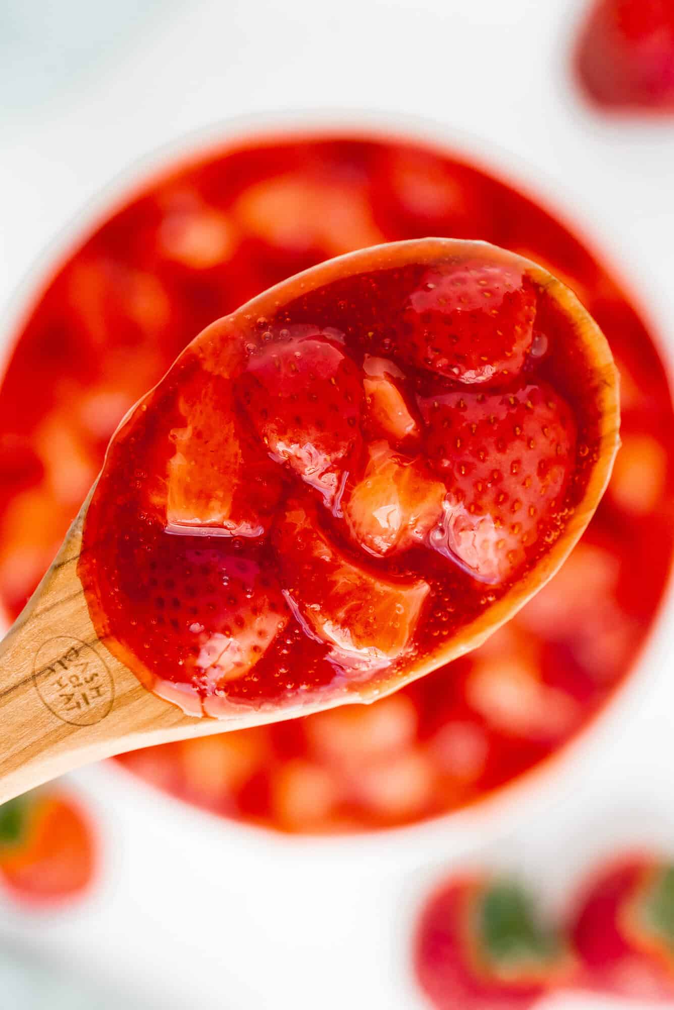 A wooden spoon holds strawberry sauce with whole and chopped strawberries in a glossy red syrup.
