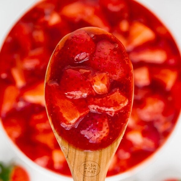 A wooden spoon holds a serving of strawberry sauce with chunks of strawberries and syrup, with a bowl of the same sauce in the background.