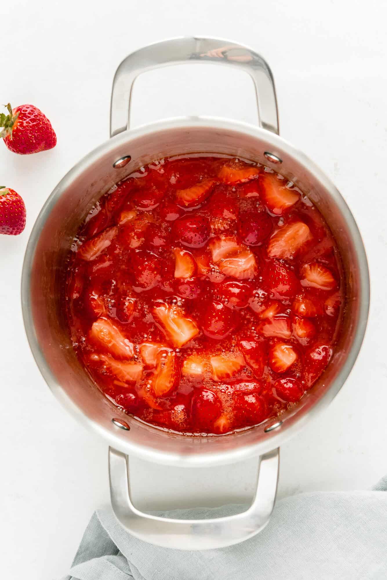 A stainless steel pot filled with simmering strawberry sauce sits on a white surface, with two whole strawberries nearby.
