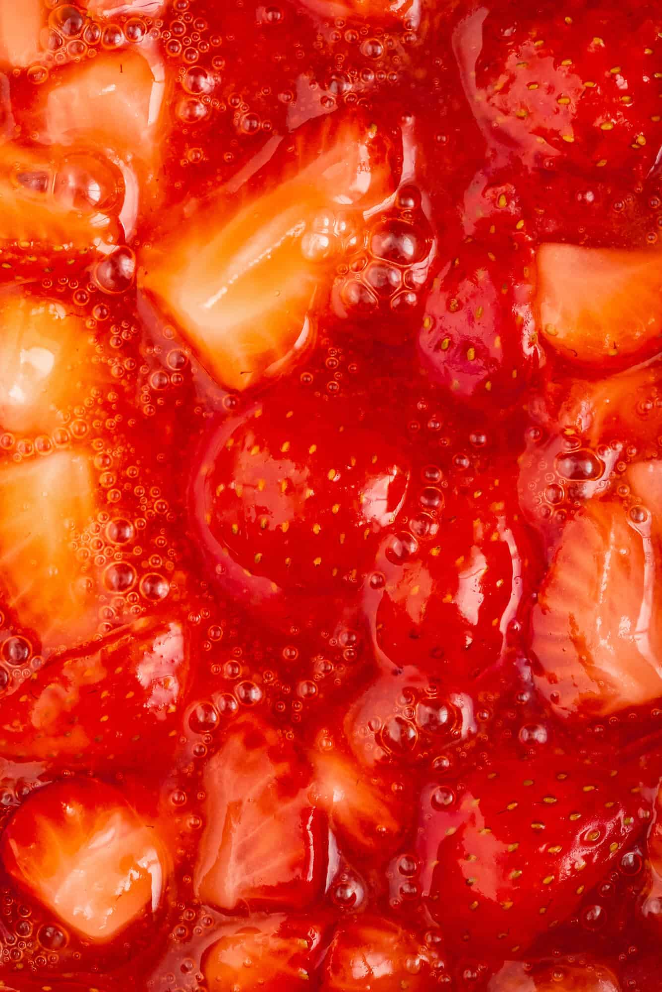 Close-up of sliced strawberries in a thick red syrup, with visible bubbles and glossy texture.