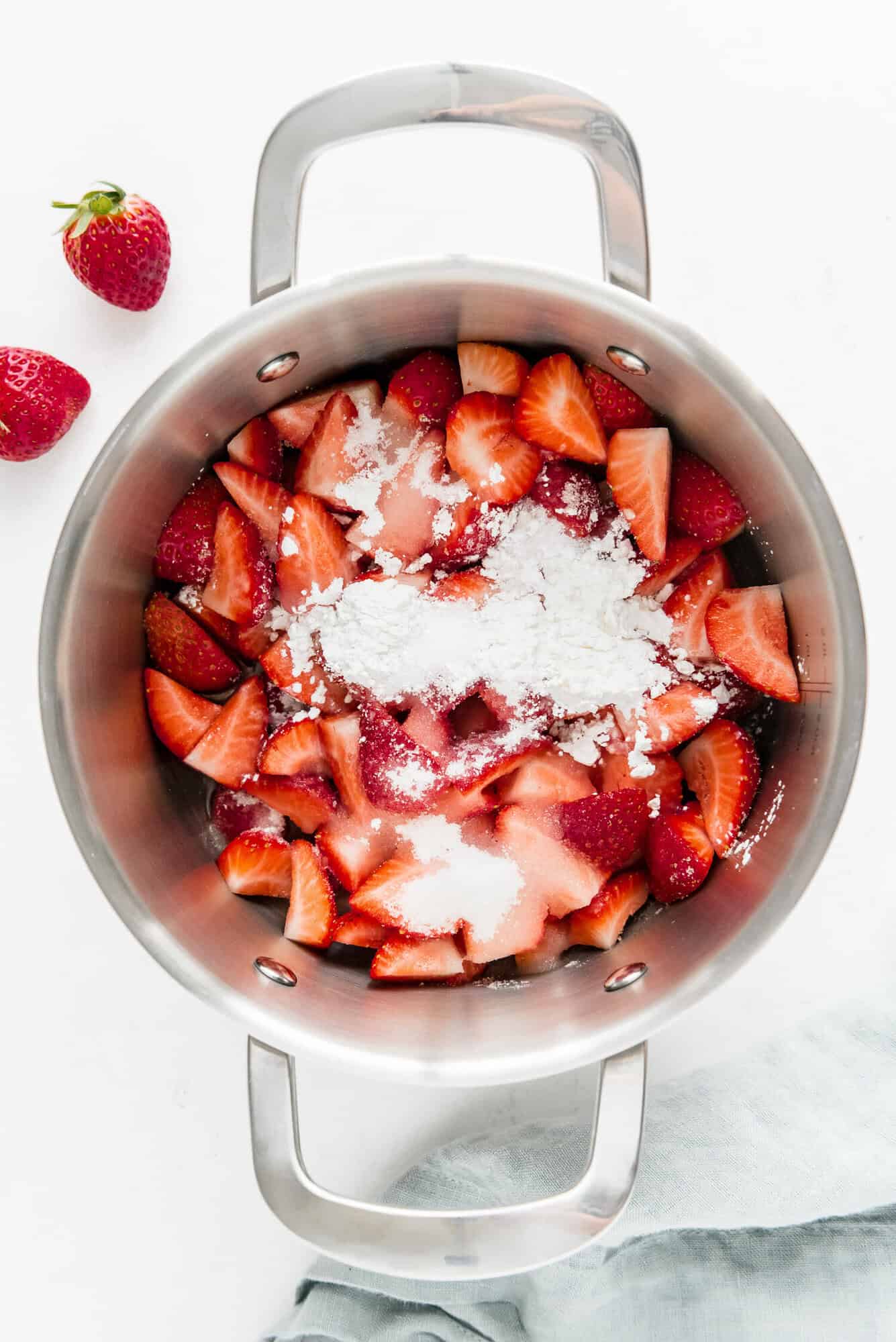 A stainless steel pot containing sliced strawberries topped with sugar and cornstarch, sits on a white surface with a few whole strawberries nearby.