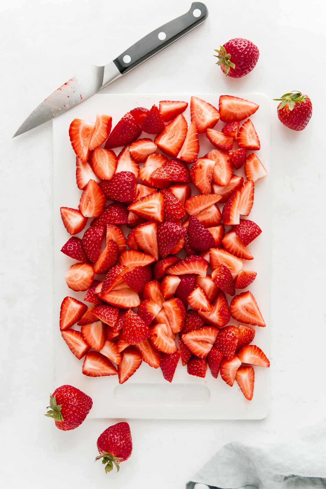 A cutting board filled with halved and quartered strawberries, a kitchen knife with strawberry residue, and whole strawberries on a white surface.