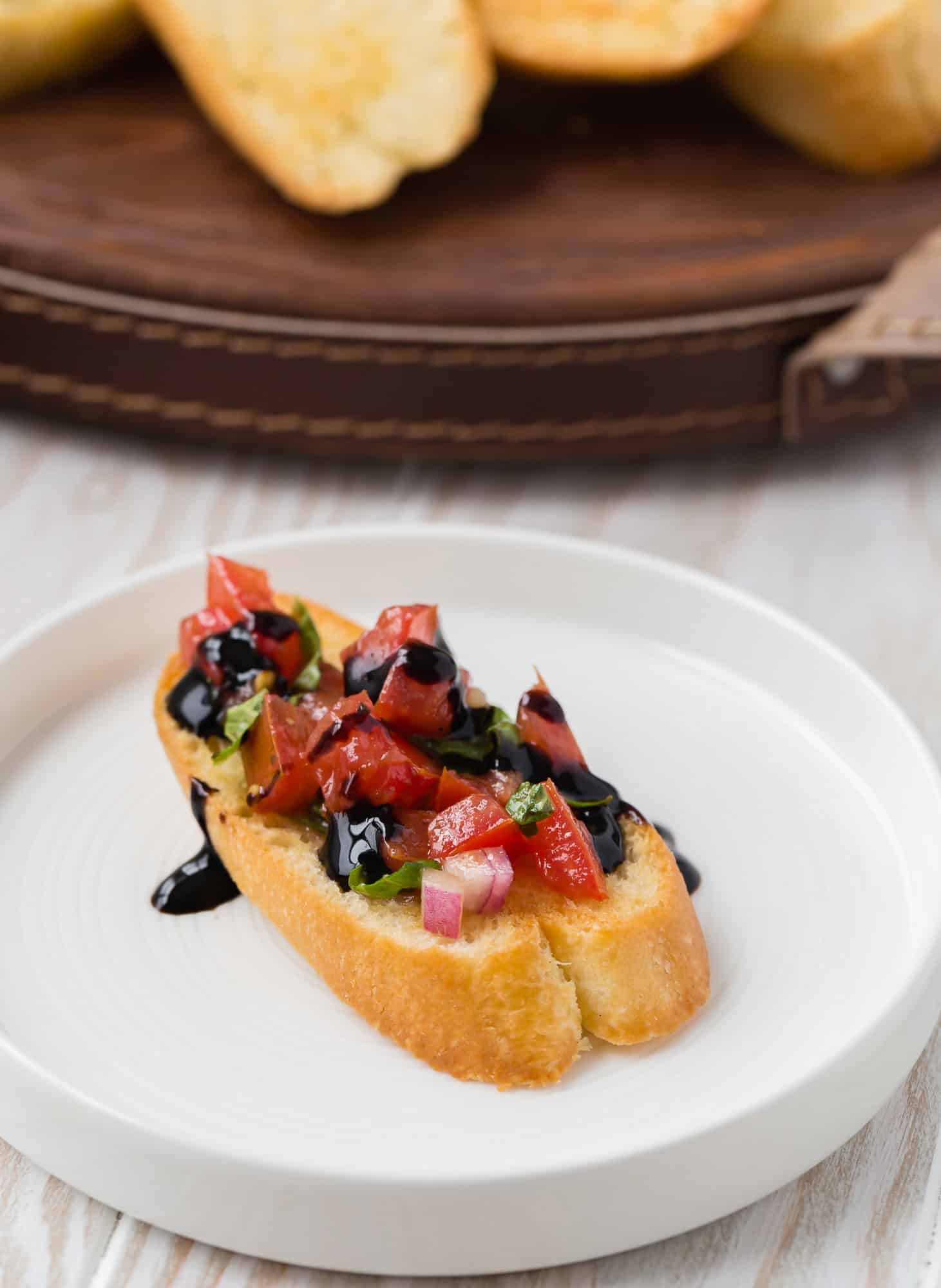 A slice of toasted bread topped with diced tomatoes, red onion, basil, and a drizzle of balsamic glaze on a white plate.