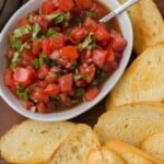 A bowl of bruschetta topping made of diced tomatoes and herbs with a spoon, surrounded by slices of toasted bread on a wooden surface.