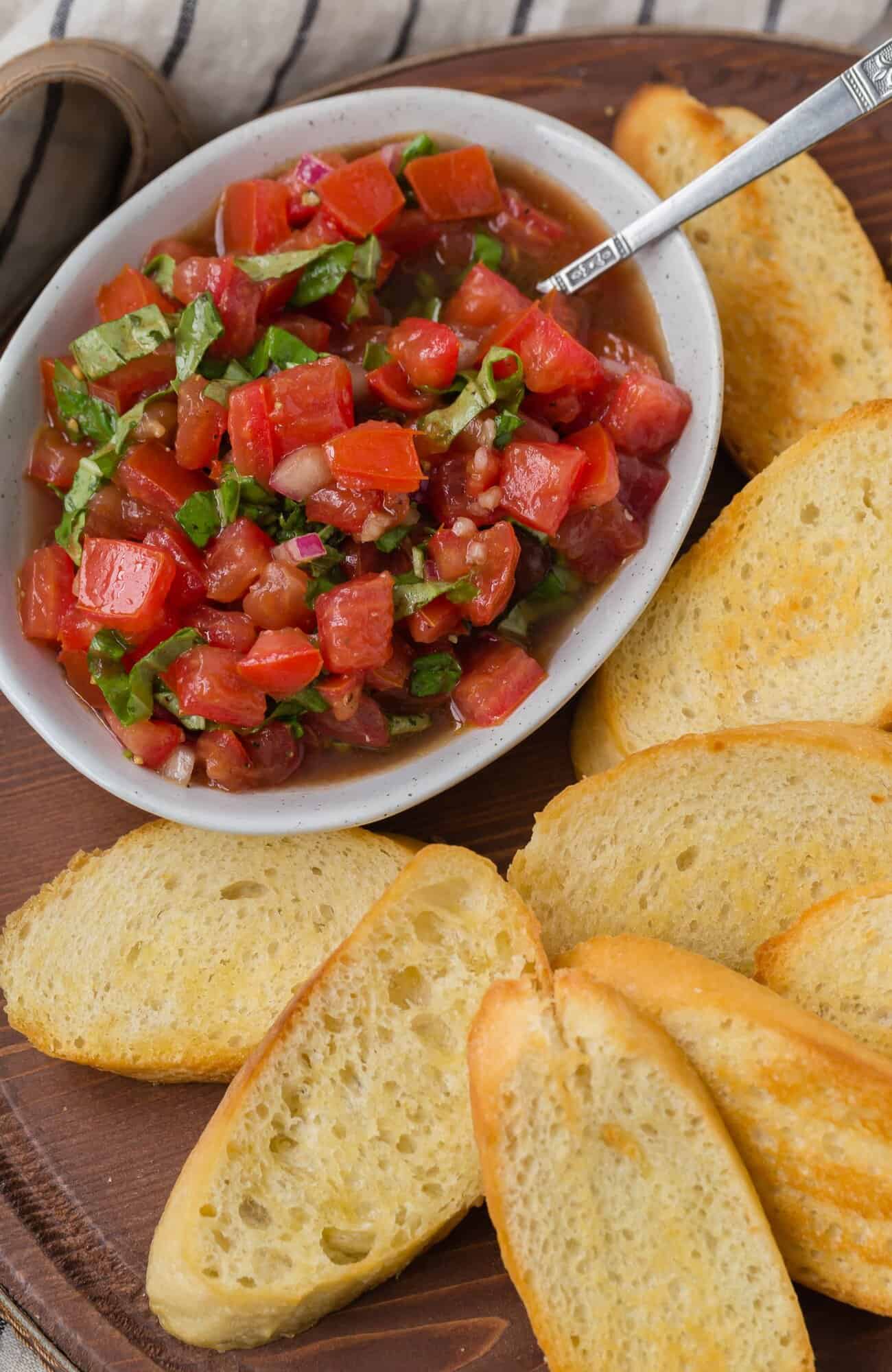 A bowl of tomato bruschetta topping with chopped tomatoes, basil, and onions beside several slices of toasted baguette on a wooden board.