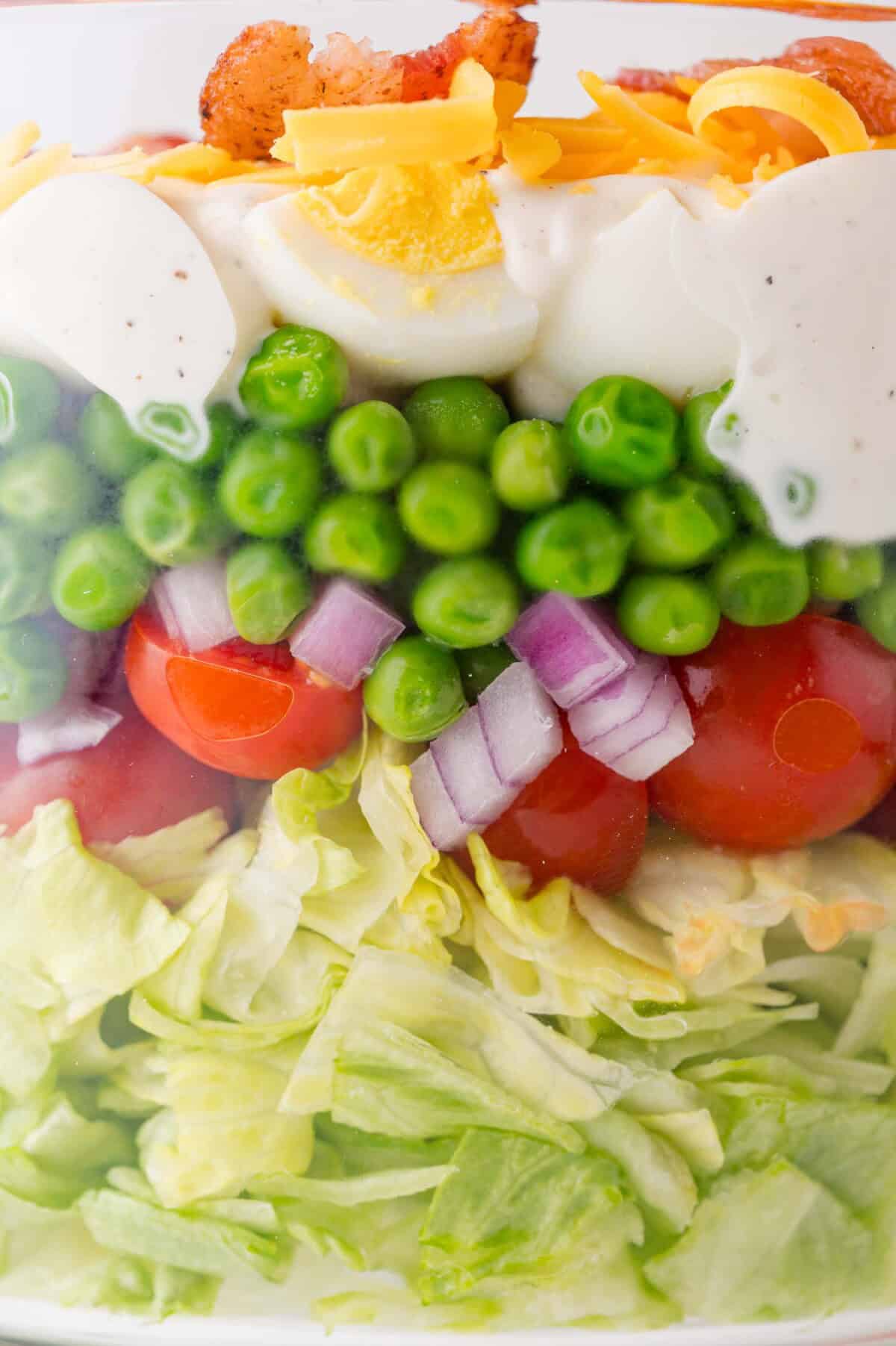 A close-up of a layered salad showing chopped lettuce, cherry tomatoes, red onions, green peas, creamy dressing, shredded cheese, and bacon.