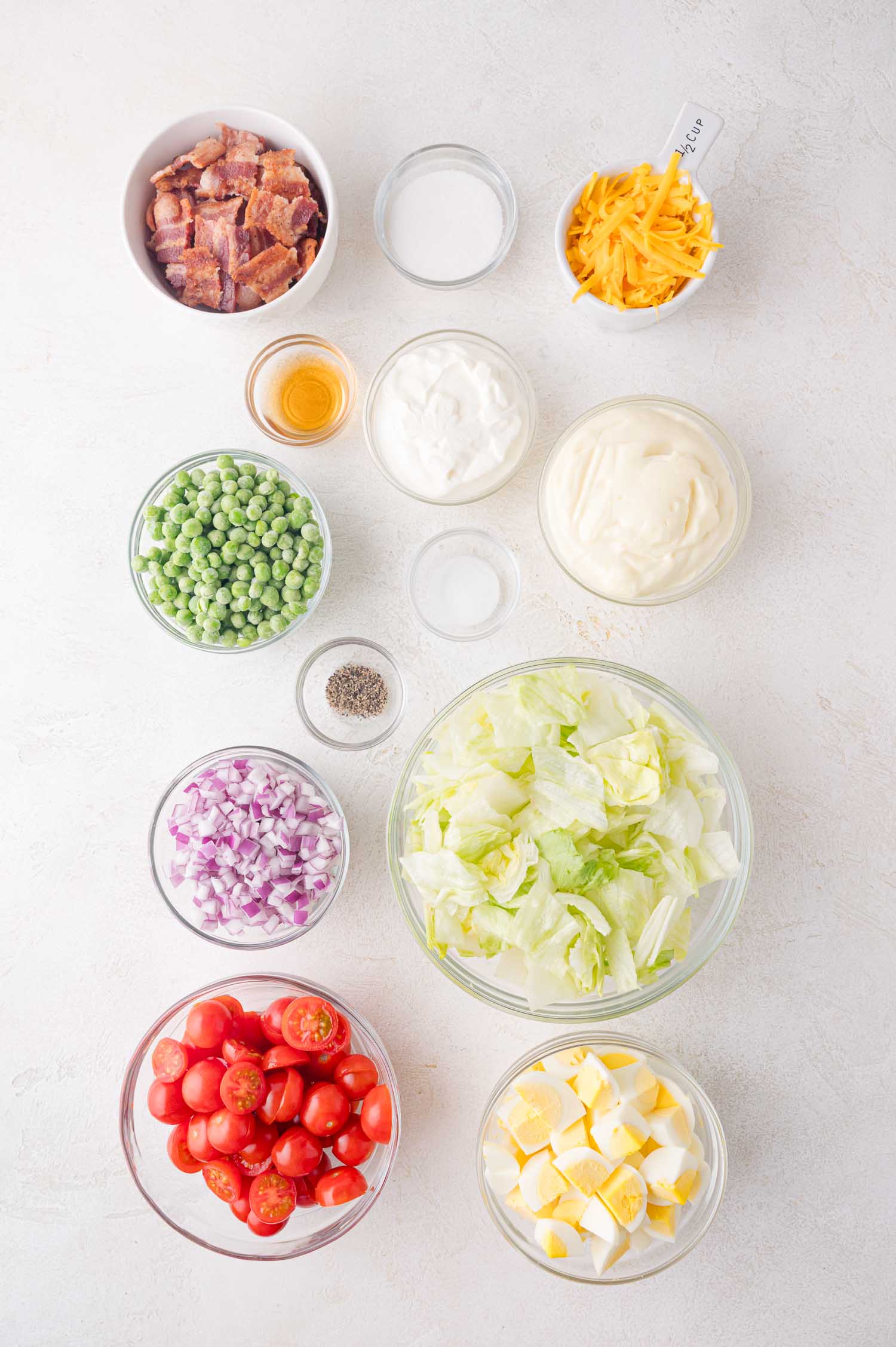 Overhead view of salad ingredients in bowls, including lettuce, cherry tomatoes, red onion, hard-boiled eggs, peas, bacon, cheese, mayonnaise, sour cream, vinegar, salt, and pepper.