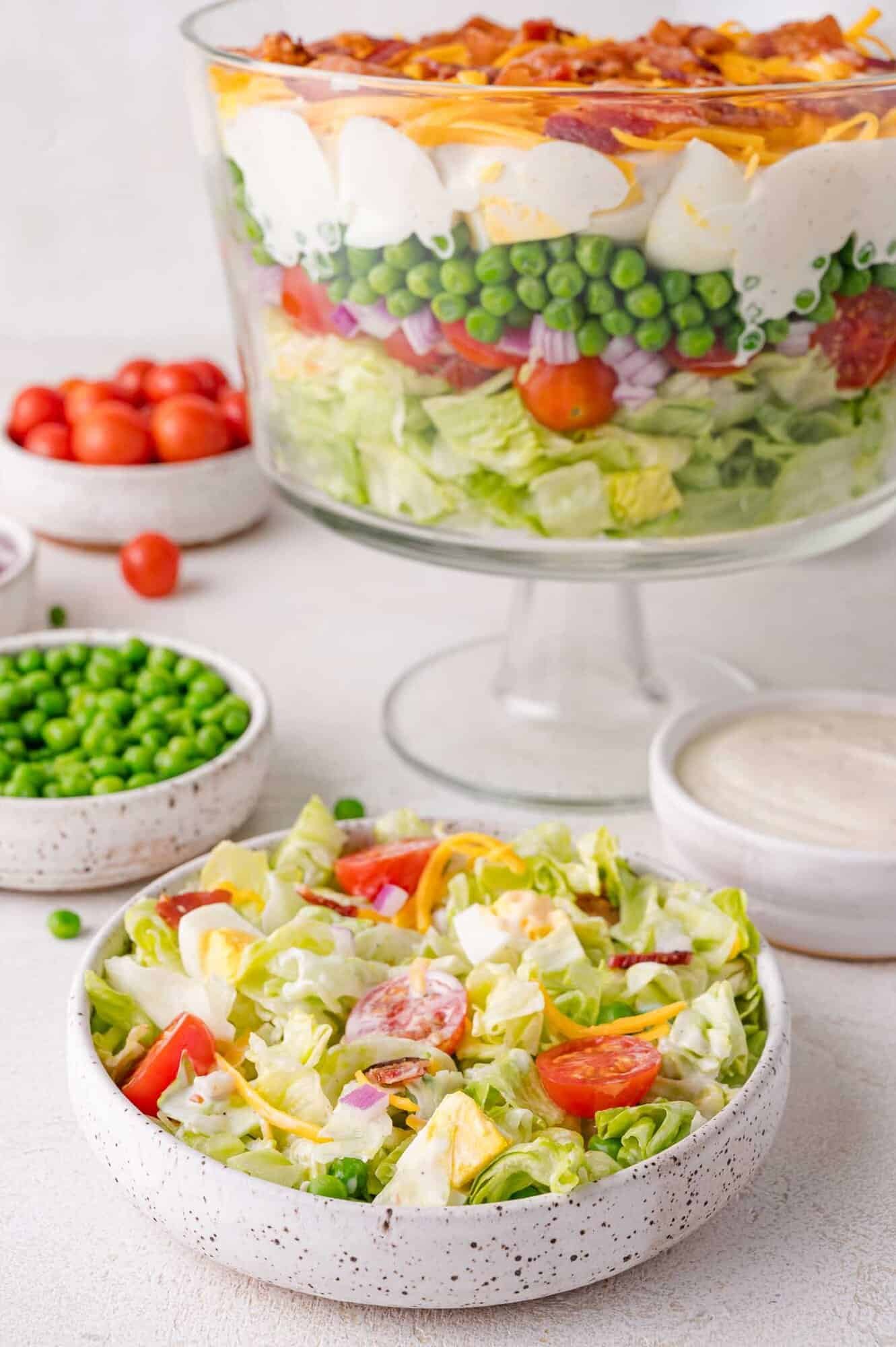 A bowl of 7 layer salad with lettuce, tomatoes, peas, eggs, cheese, and bacon next to a small bowl of salad, dressing, peas, and cherry tomatoes.