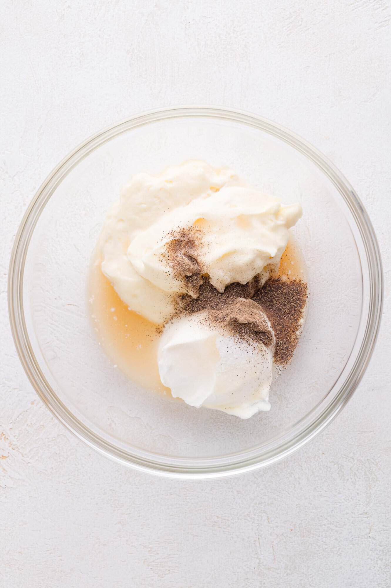 A glass bowl containing mayonnaise, sour cream, vinegar, sugar, salt, and pepper on a white surface.