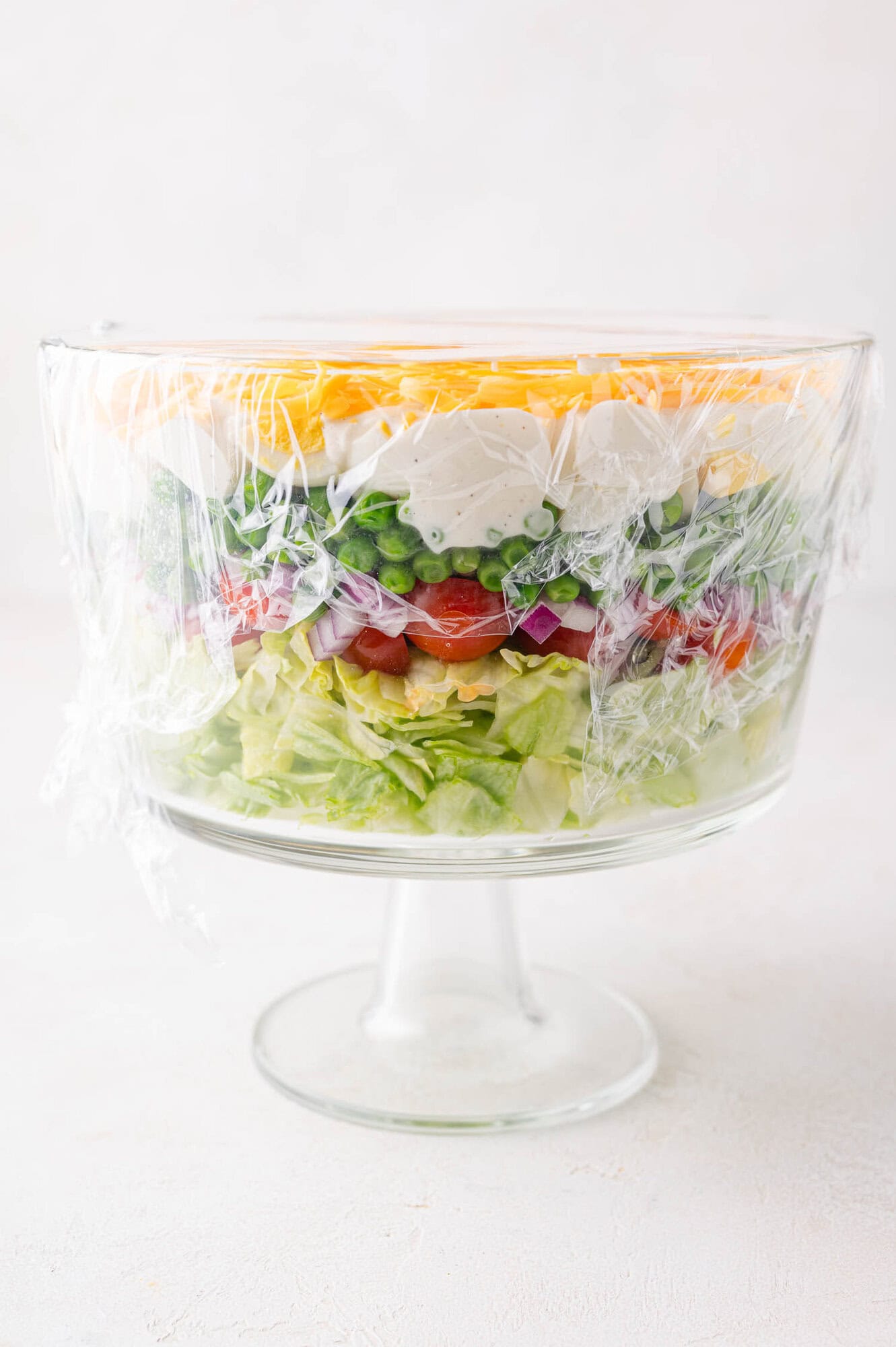 A glass trifle bowl filled with neatly layered salad ingredients, covered in plastic wrap.