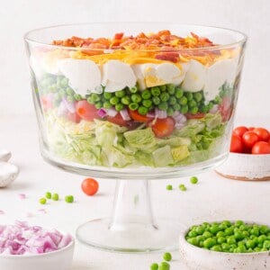 A glass trifle bowl filled with layered salad ingredients including lettuce, tomatoes, red onion, peas, eggs, cheese, and bacon.