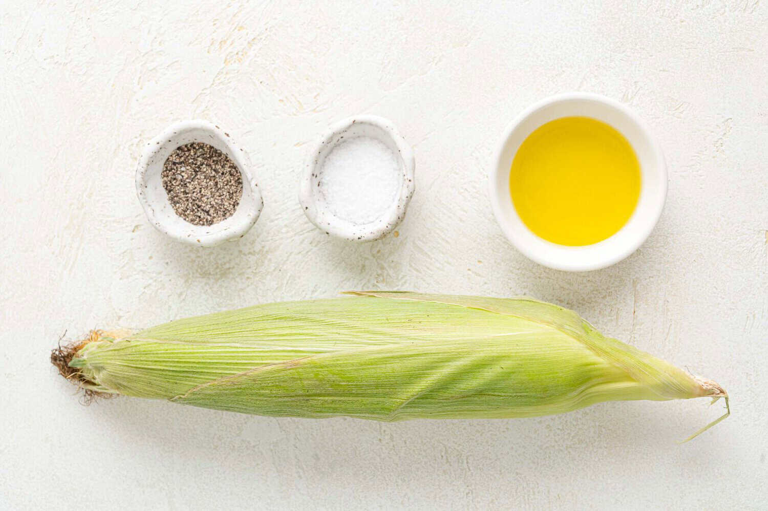An ear of corn in husk is placed on a white surface with small bowls of black pepper, salt, and olive oil arranged above it.