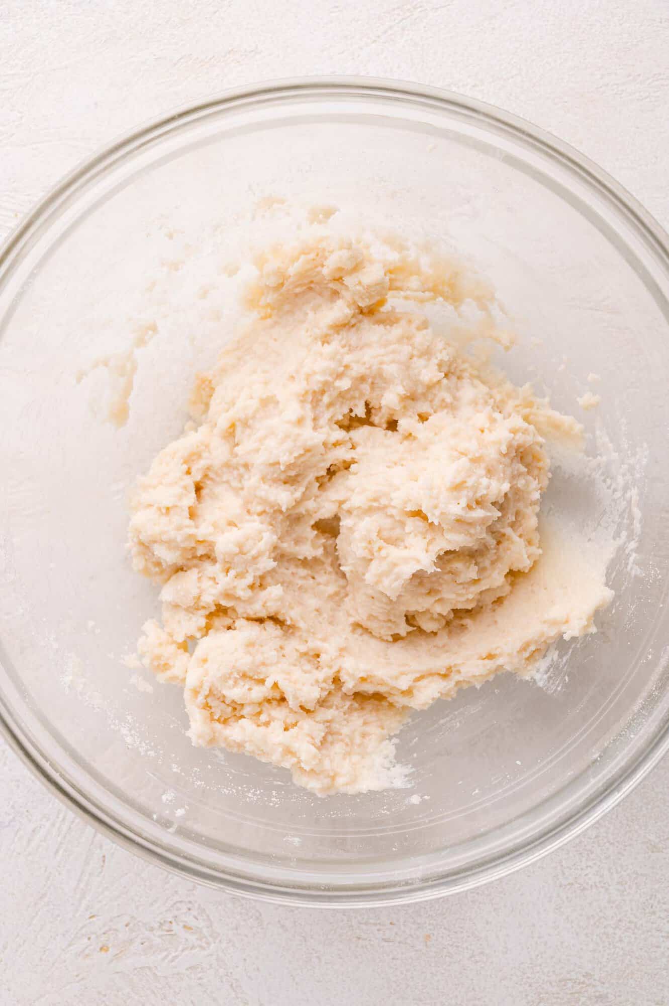 A clear glass bowl containing a mixture of pale, thick dough on a light-colored surface.