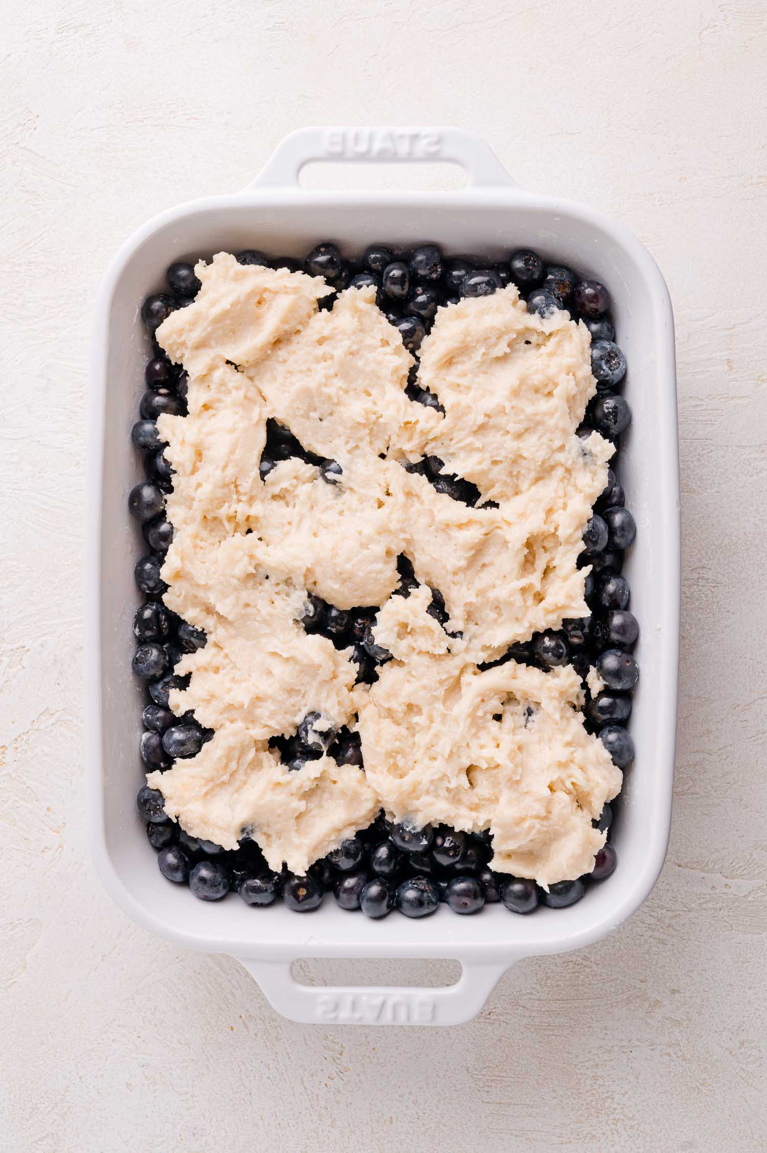 A white baking dish filled with fresh blueberries topped with uneven dollops of biscuit dough, ready to be baked.