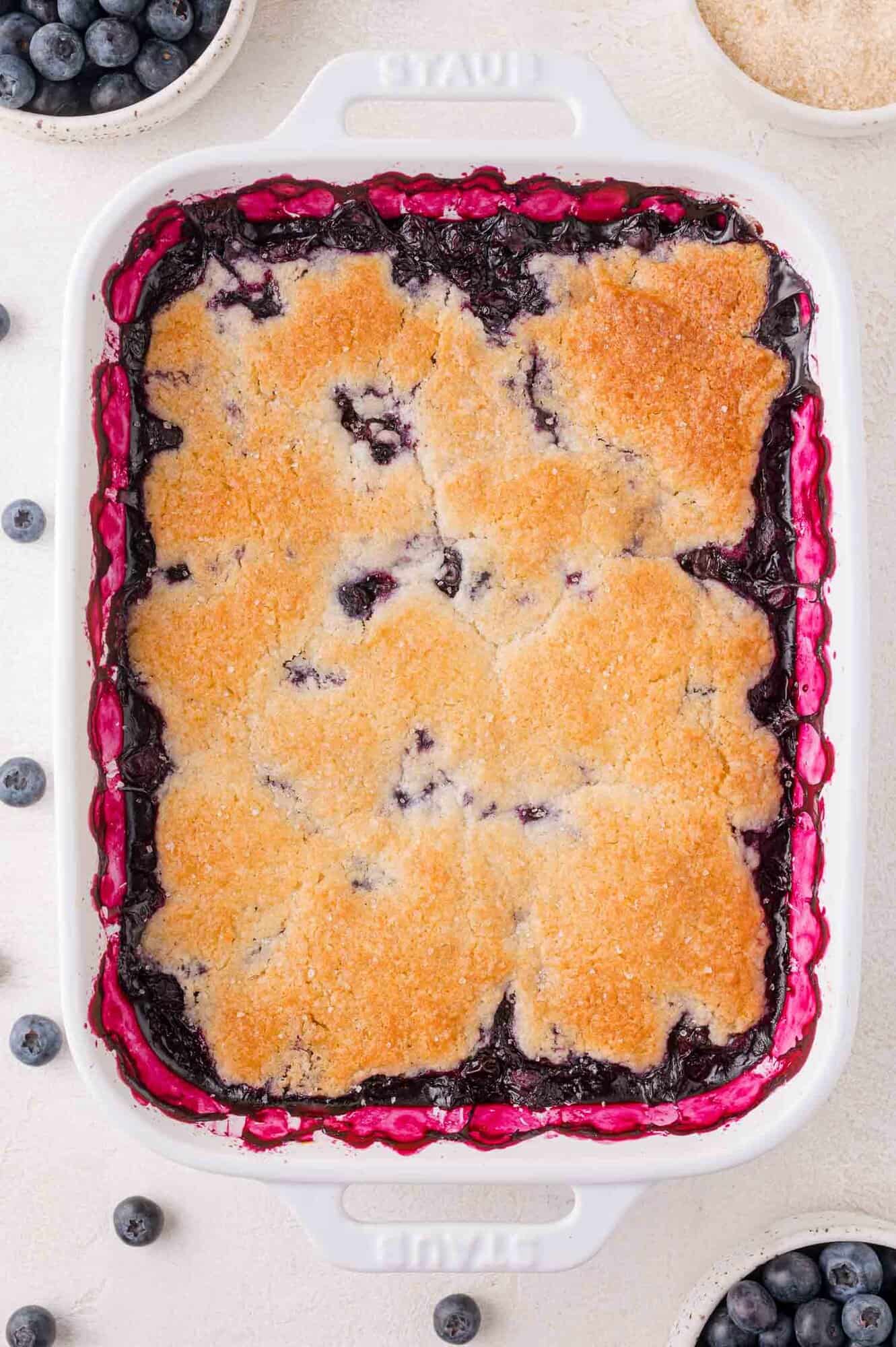 A baked blueberry cobbler with a golden-brown crust in a white rectangular dish, surrounded by fresh blueberries and small bowls of ingredients.