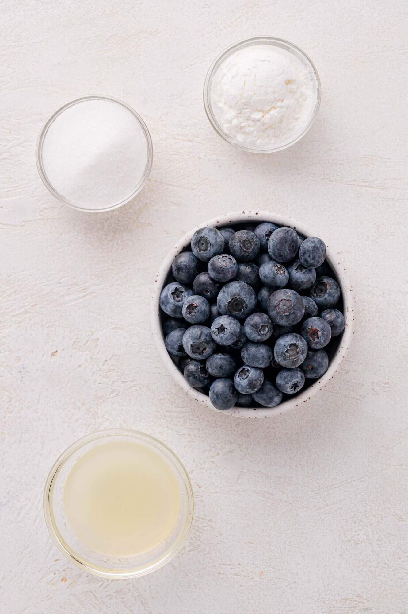 A bowl of blueberries, a small bowl of sugar, a small bowl of cornstarch, and a small cup of lemon juice on a light surface.
