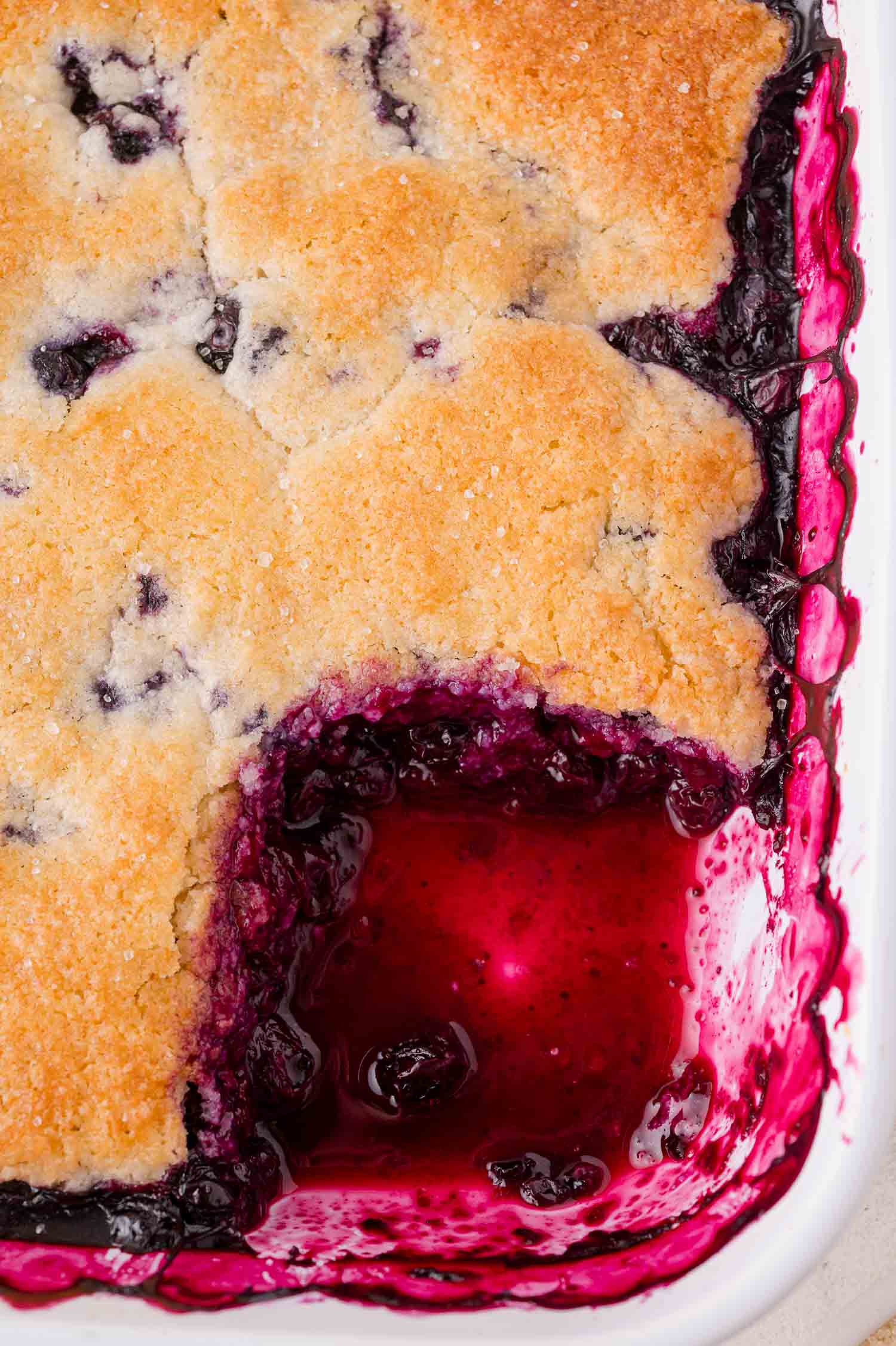A close-up of a baked blueberry cobbler in a white dish with a portion scooped out, revealing the dark purple fruit filling beneath a golden-brown crust.