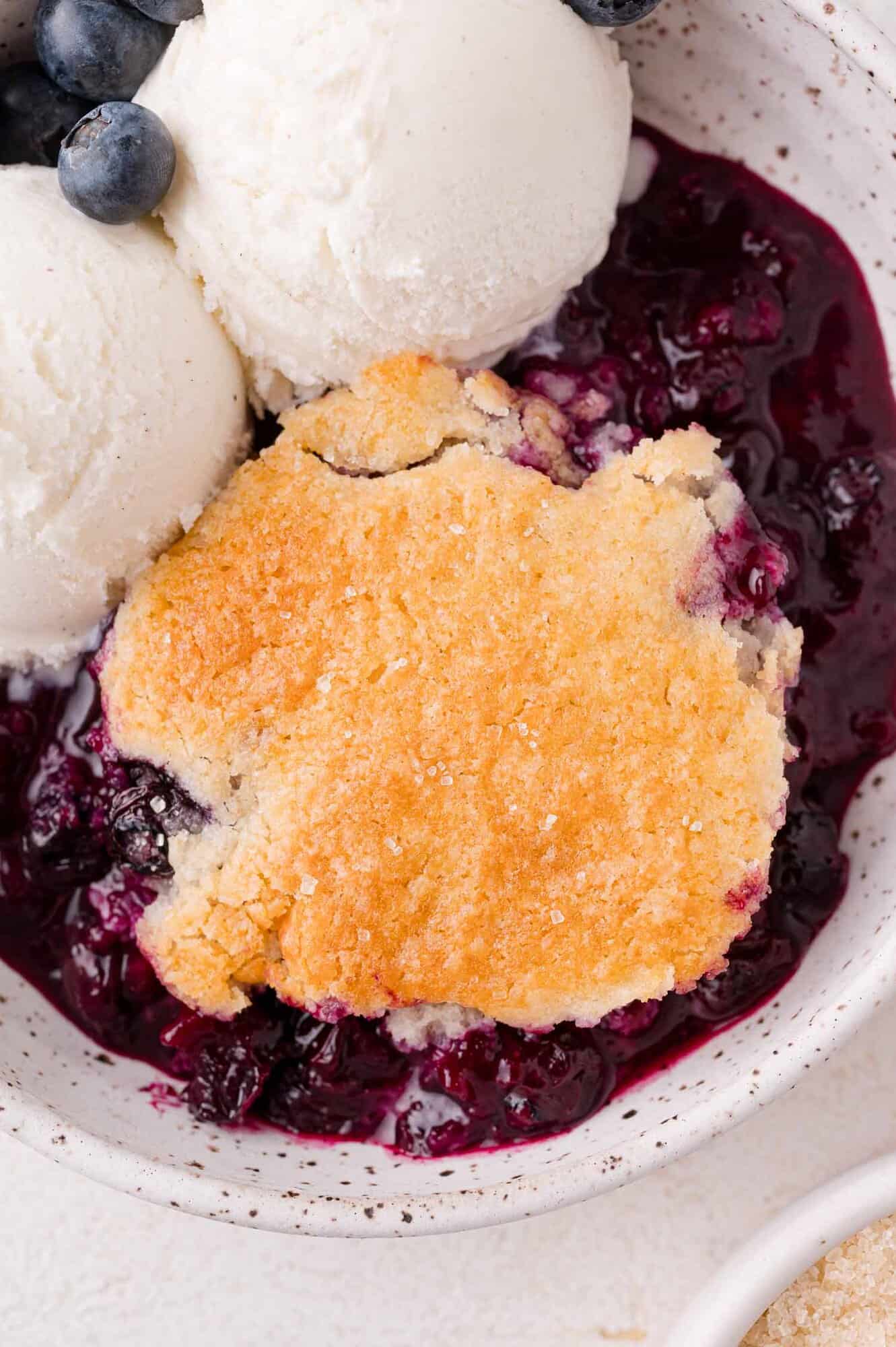 A serving of blueberry cobbler with a golden crust, topped with two scoops of vanilla ice cream and garnished with fresh blueberries in a white bowl.