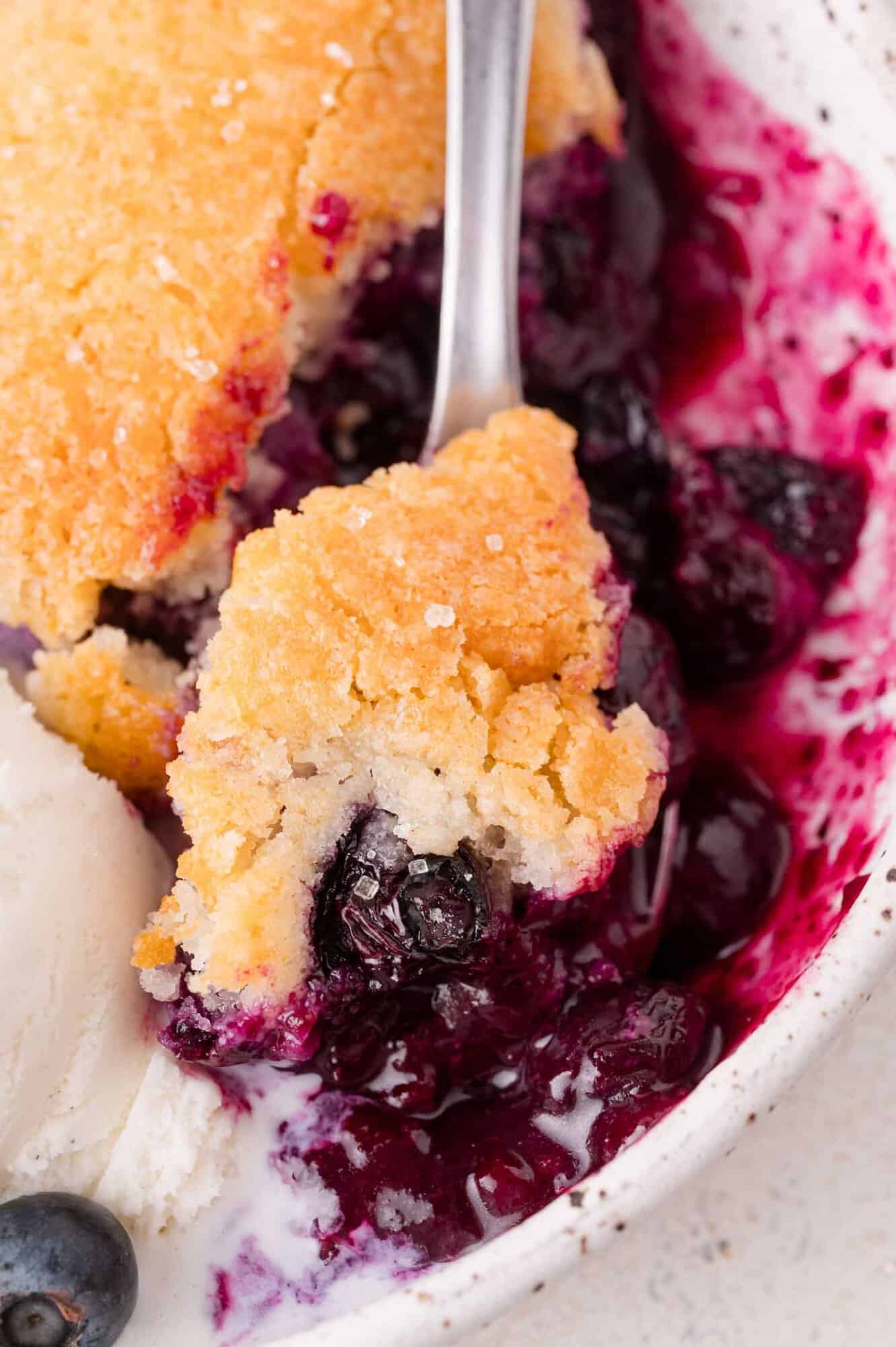 A close-up of a blueberry cobbler with a golden biscuit topping, a spoonful lifted, served with a scoop of vanilla ice cream on the side.