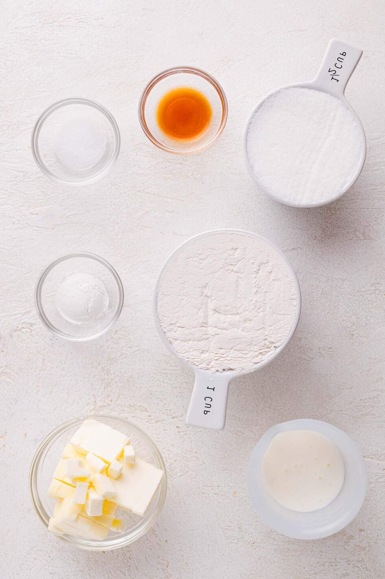 Assorted baking ingredients including flour, sugar, butter, buttermilk, vanilla extract, baking powder, and salt arranged in bowls on a white surface.