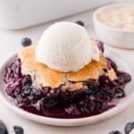 A serving of blueberry cobbler topped with a scoop of vanilla ice cream on a white plate, with fresh blueberries visible nearby.