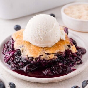 A serving of blueberry cobbler topped with a scoop of vanilla ice cream on a white plate, with fresh blueberries visible nearby.