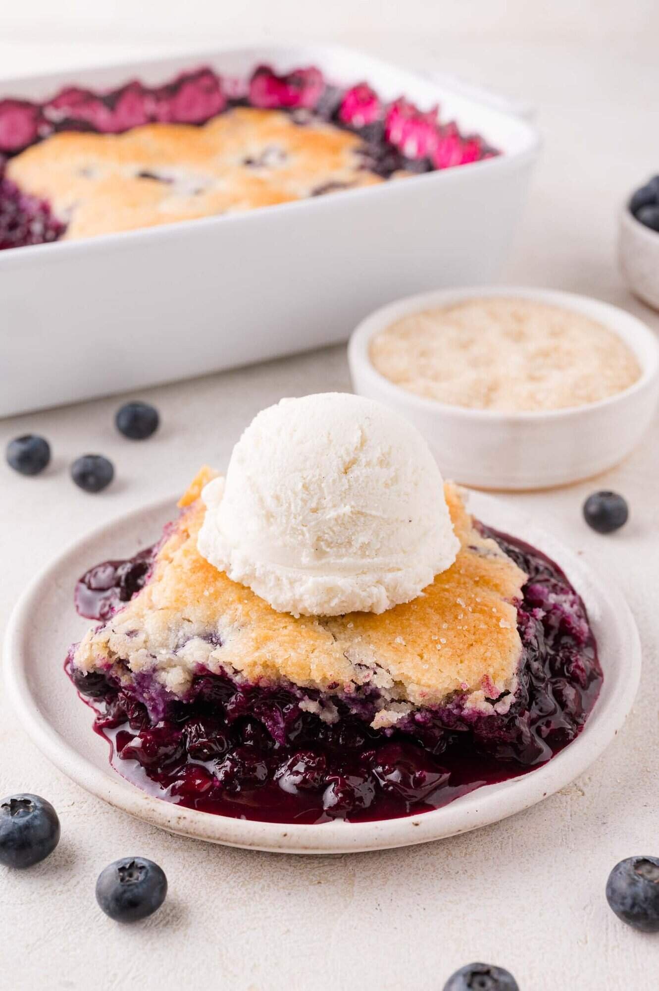 A slice of blueberry cobbler topped with a scoop of vanilla ice cream on a plate, with fresh blueberries and a dish of sugar in the background.