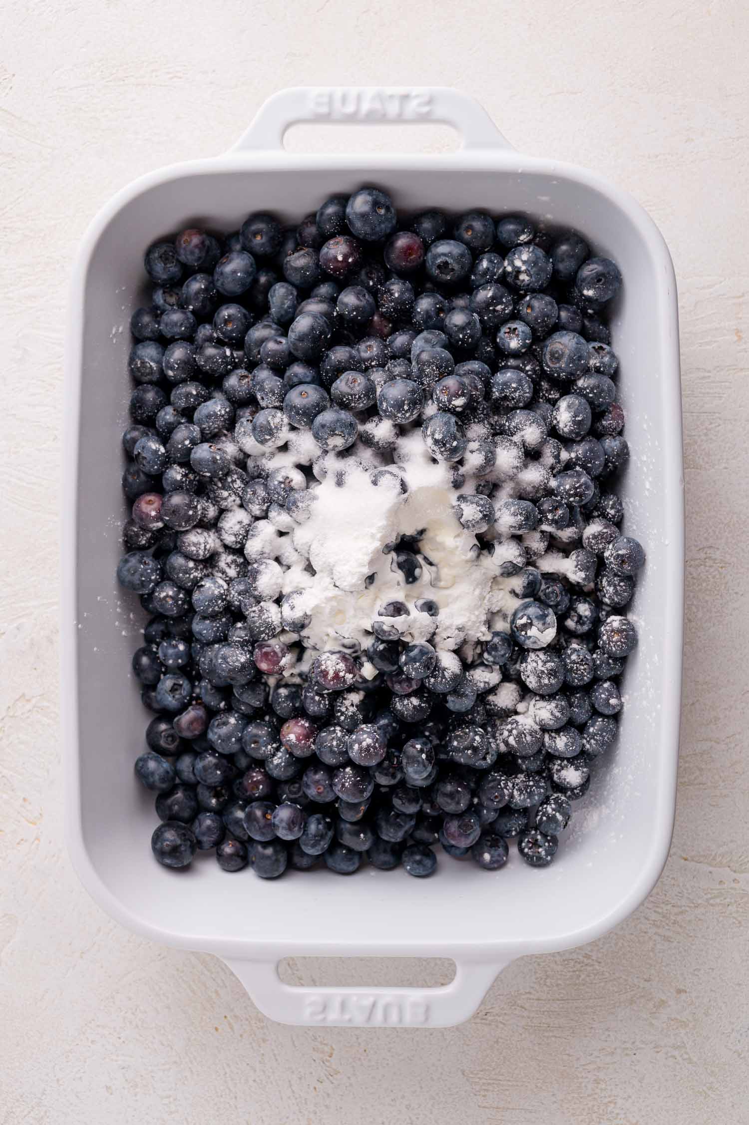 A white baking dish filled with fresh blueberries topped with a pile of sugar and cornstarch.