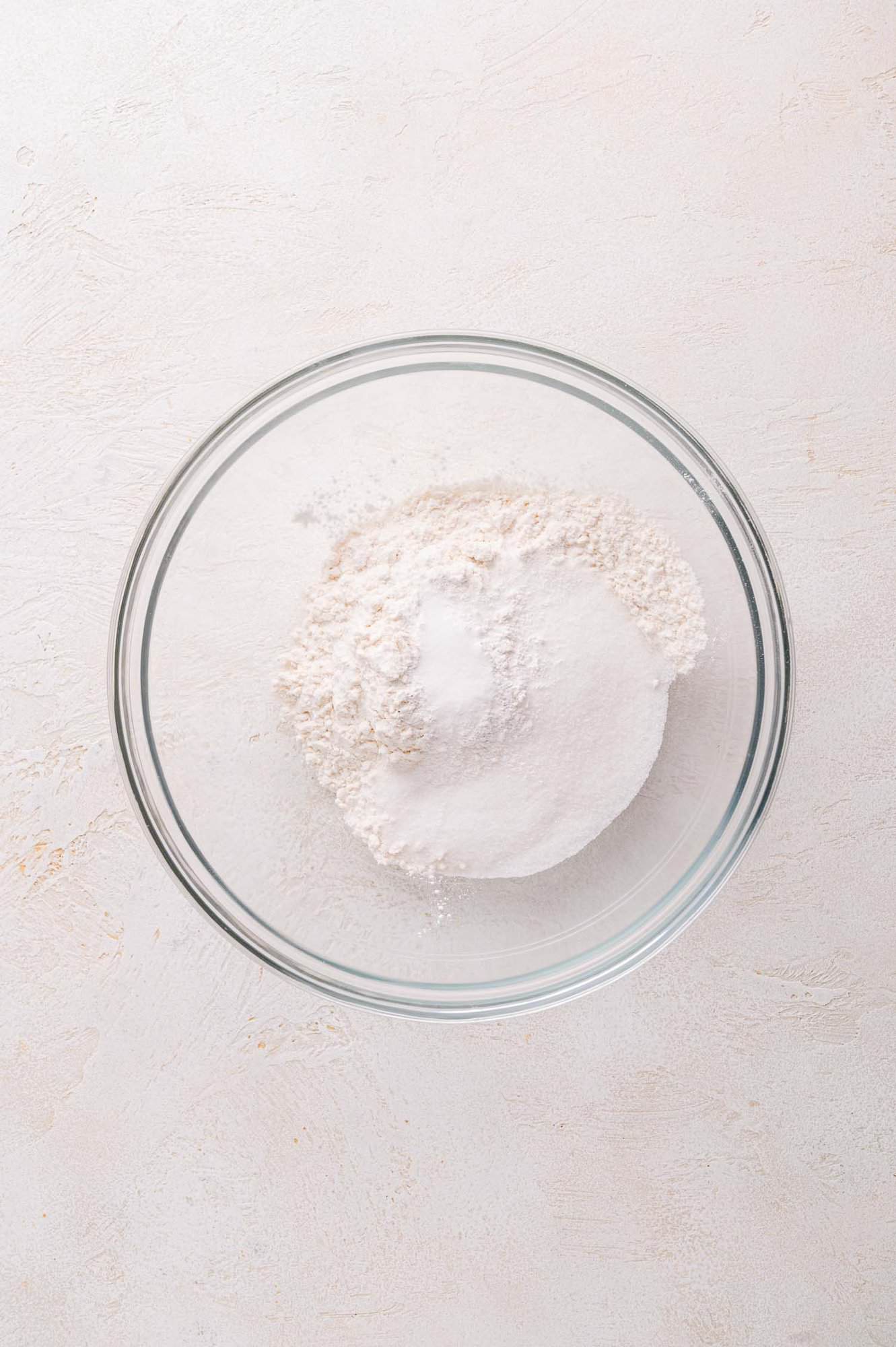 A clear glass bowl containing a mound of white flour and a small amount of granulated sugar, set on a light textured surface.