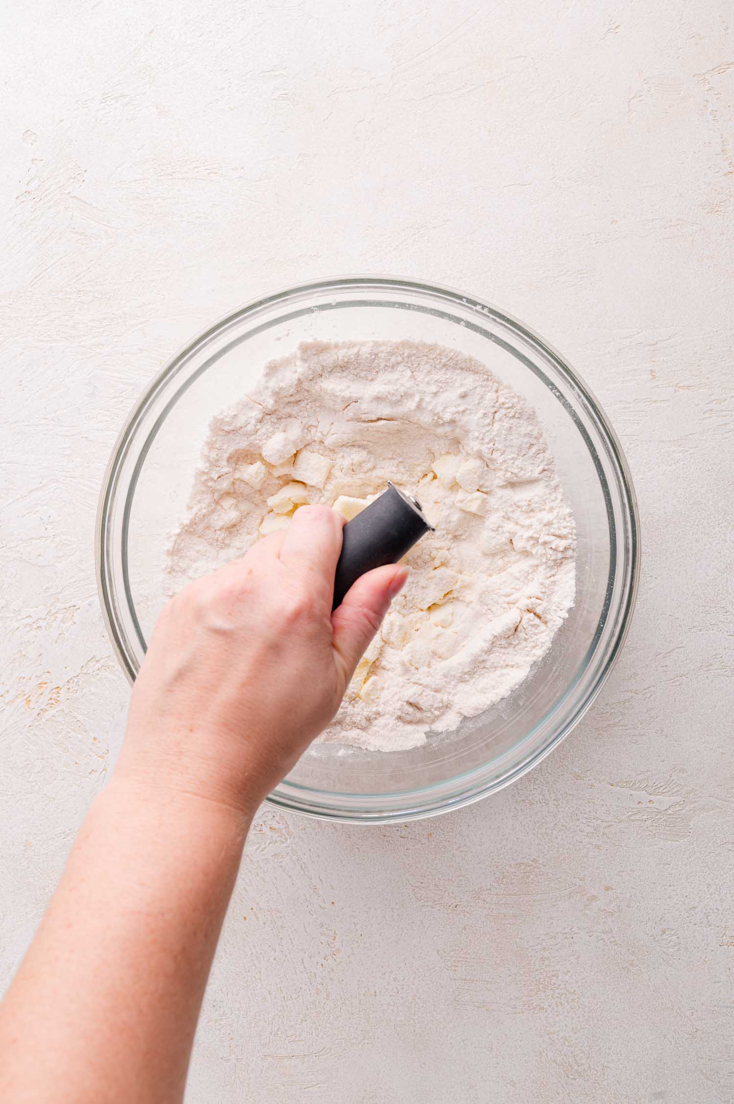 A hand uses a pastry cuttter to cut butter into flour in a glass mixing bowl on a light-colored surface.