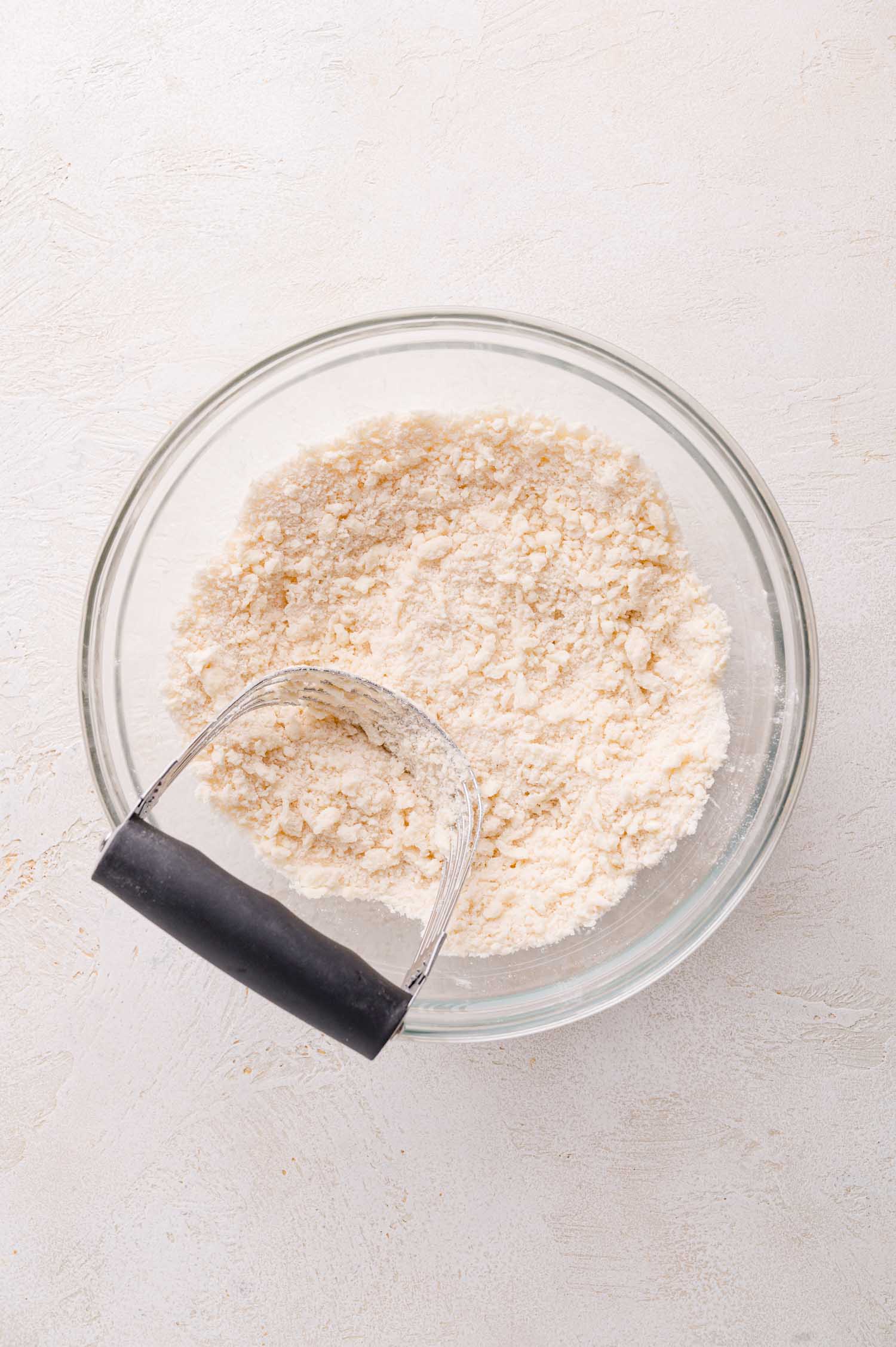 A glass bowl contains a flour mixture with a pastry cutter resting inside, set on a light-colored surface.