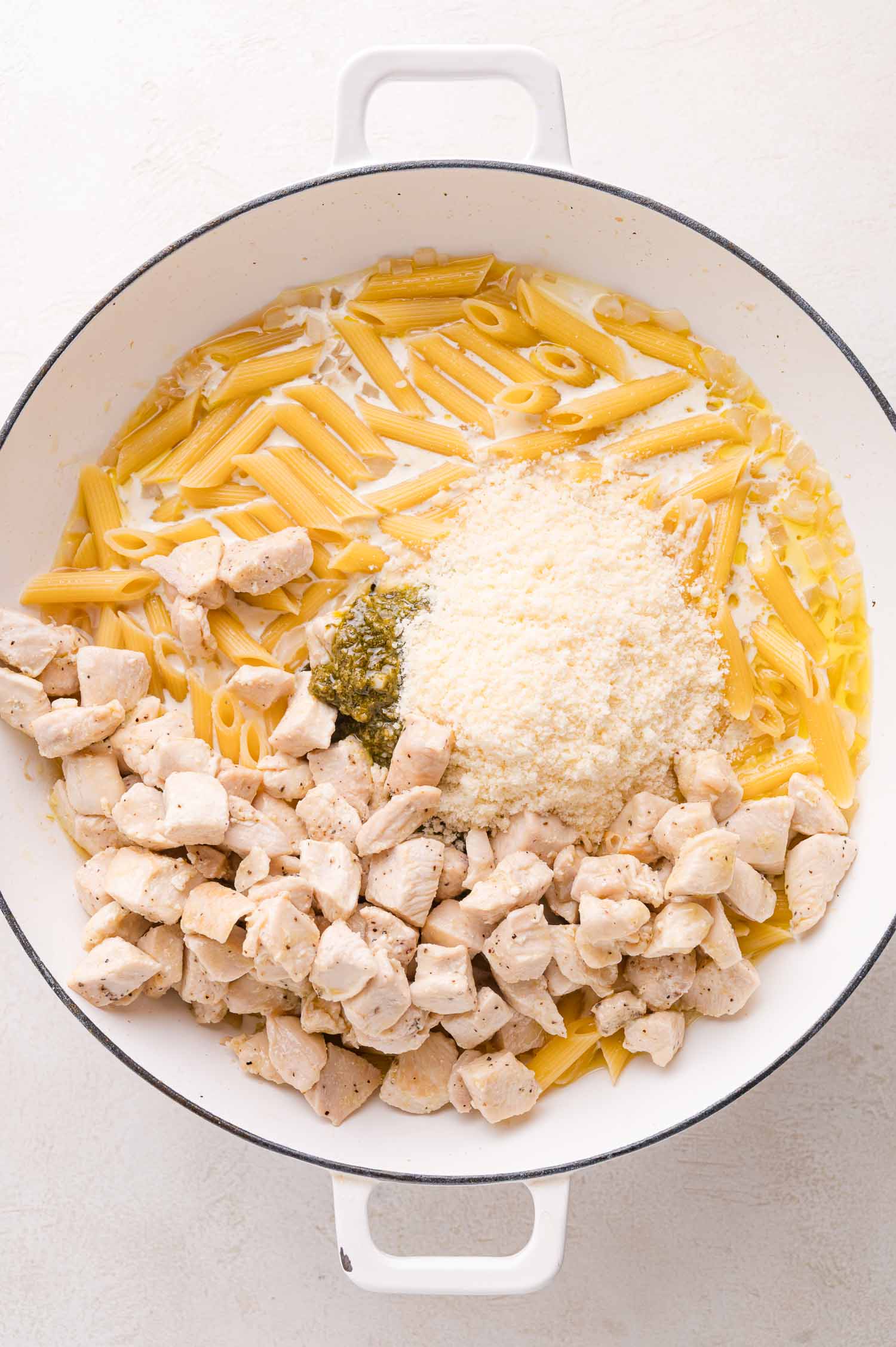 A white pan containing uncooked penne pasta, cooked diced chicken, pesto sauce, grated parmesan cheese, and liquid ingredients.