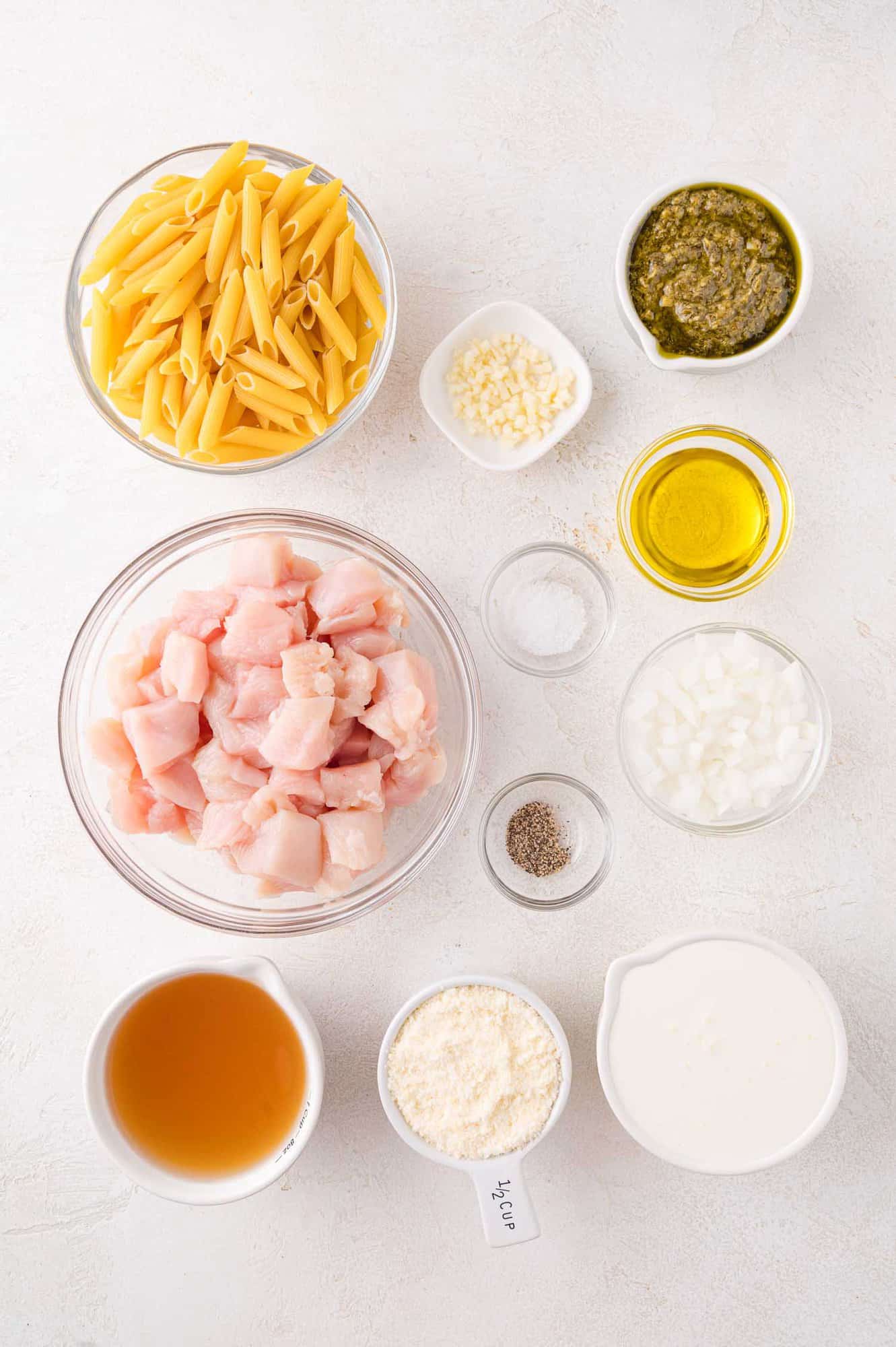 Ingredients for a pasta dish arranged on a white surface, including penne, diced raw chicken, pesto, olive oil, cream, grated cheese, chopped onion, minced garlic, broth, salt, and pepper.