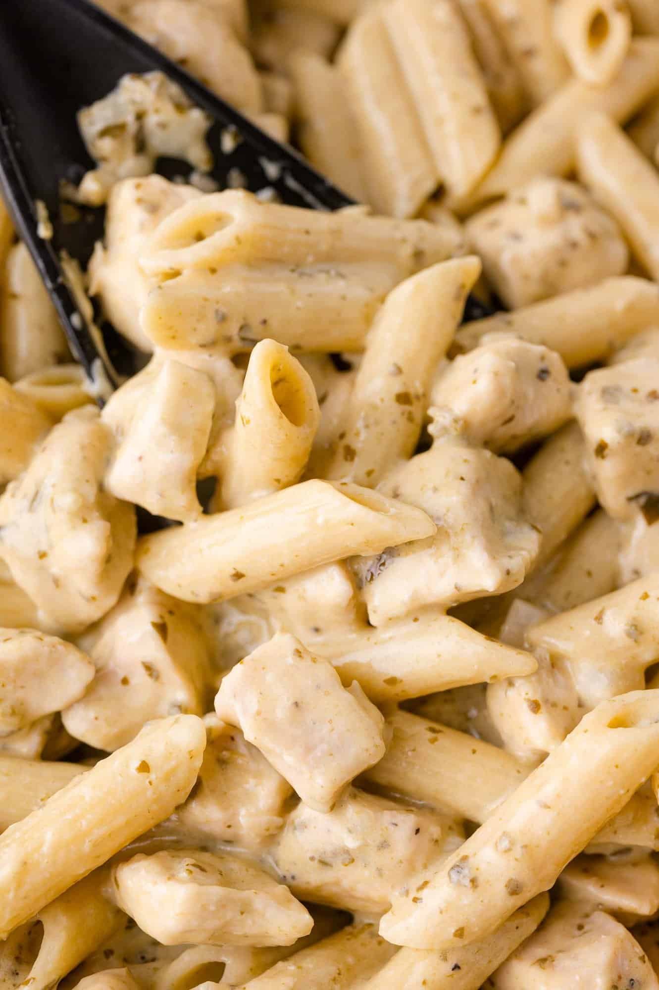 Close-up of creamy pesto chicken pasta with creamy white sauce, herbs, and chunks of chicken, with a black spoon mixing the dish.