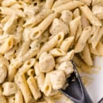 Close-up of creamy penne pasta with pieces of chicken, mixed with a light sauce and herbs, being stirred with a black serving spoon.